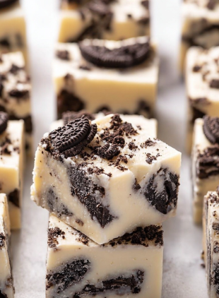 The image shows a close-up of thick square pieces of cookies and cream fudge layered in white creamy base mixed with broken chocolate cookie bits. Each piece has a smooth, pale off-white layer filled with dark brown cookie chunks inside and baked bits scattered on top, creating a textured look with irregular shapes. The fudge pieces are placed closely together on a white marbled surface, highlighting the contrast between the creamy white fudge and dark cookies. Photo taken with an iphone --ar 2:3 --v 7 - White Chocolate Oreo Fudge, Oreo fudge recipe, no-bake Oreo fudge, creamy white chocolate fudge, easy Oreo dessert