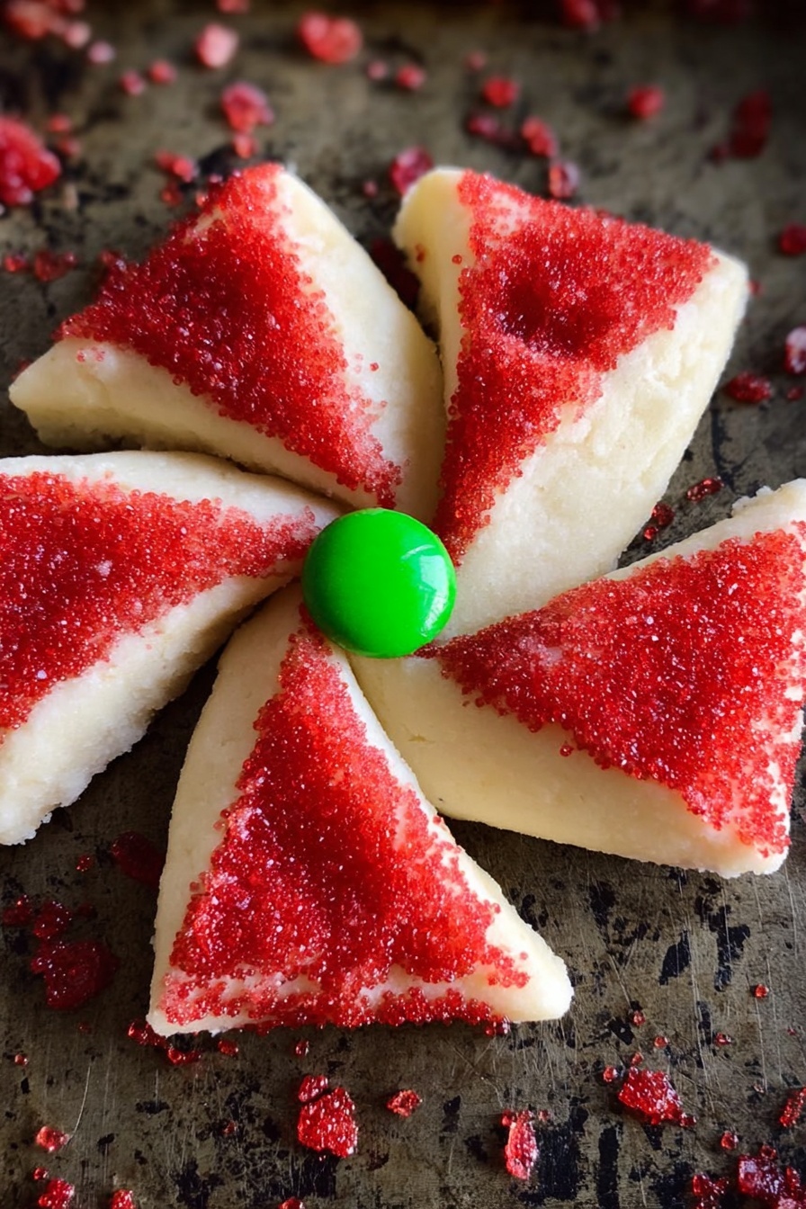 The image shows a pinwheel-shaped cookie with four red sugar-coated triangular sections alternating with four plain white dough triangular sections folded over slightly. A single bright green round candy is placed at the center where all four triangles meet. The cookie sits on a dark baking tray sprinkled with scattered red sugar crystals. The white dough looks smooth and creamy, while the red sugar sprinkles add a rough texture. Photo taken with an iphone --ar 2:3 --v 7 - Poinsettia Cookies with M&Ms, holiday cookie ideas, festive sugar cookies, flower-shaped Christmas cookies, easy holiday cookie recipes