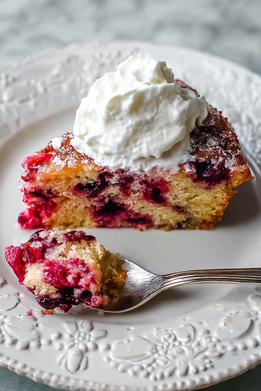 A slice of berry cake is placed on a white decorative plate with raised floral patterns. The cake has two visible layers: the bottom layer is a light golden-brown sponge filled with bright red and dark purple berries throughout, while the top layer appears slightly caramelized with a glossy texture and scattered sugar crystals. A generous dollop of fluffy white whipped cream sits on top of the cake. To the left of the cake, a silver spoon holds a small bite of the berry cake, showing its moist and chunky berry-filled texture. The plate rests on a white marbled surface photo taken with an iphone --ar 2:3 --v 7 - Cranberry Pie with Nuts, Cranberry Pie, Nutty Cranberry Tart, Holiday Cranberry Dessert, Tart and Nutty Cranberry Pie