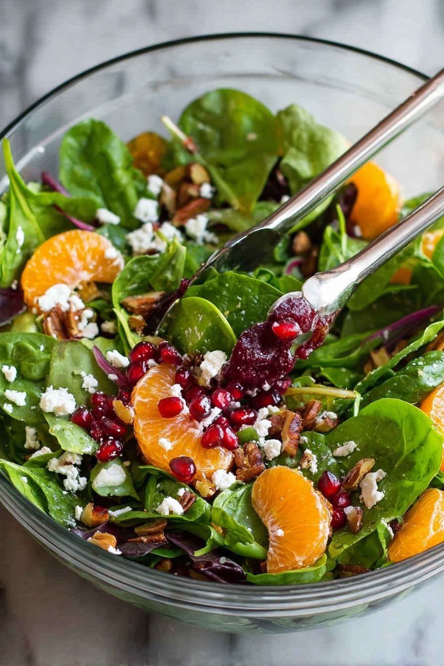 A clear glass bowl holds a fresh salad with multiple layers: the bottom layer is bright green spinach and mixed leafy greens, scattered with small white cheese crumbles. On top, there are round, orange mandarin slices and deep red pomegranate seeds mixed throughout. Some brown pieces of nuts are spread across the salad, adding texture. Silver salad tongs are partially sunk into the salad, holding some greens and a mandarin slice. The bowl sits on a white marbled surface. photo taken with an iphone --ar 2:3 --v 7 - Festive Citrus Pomegranate Salad, holiday citrus salad, easy holiday fruit salad, healthy festive salad, Christmas citrus fruit salad
