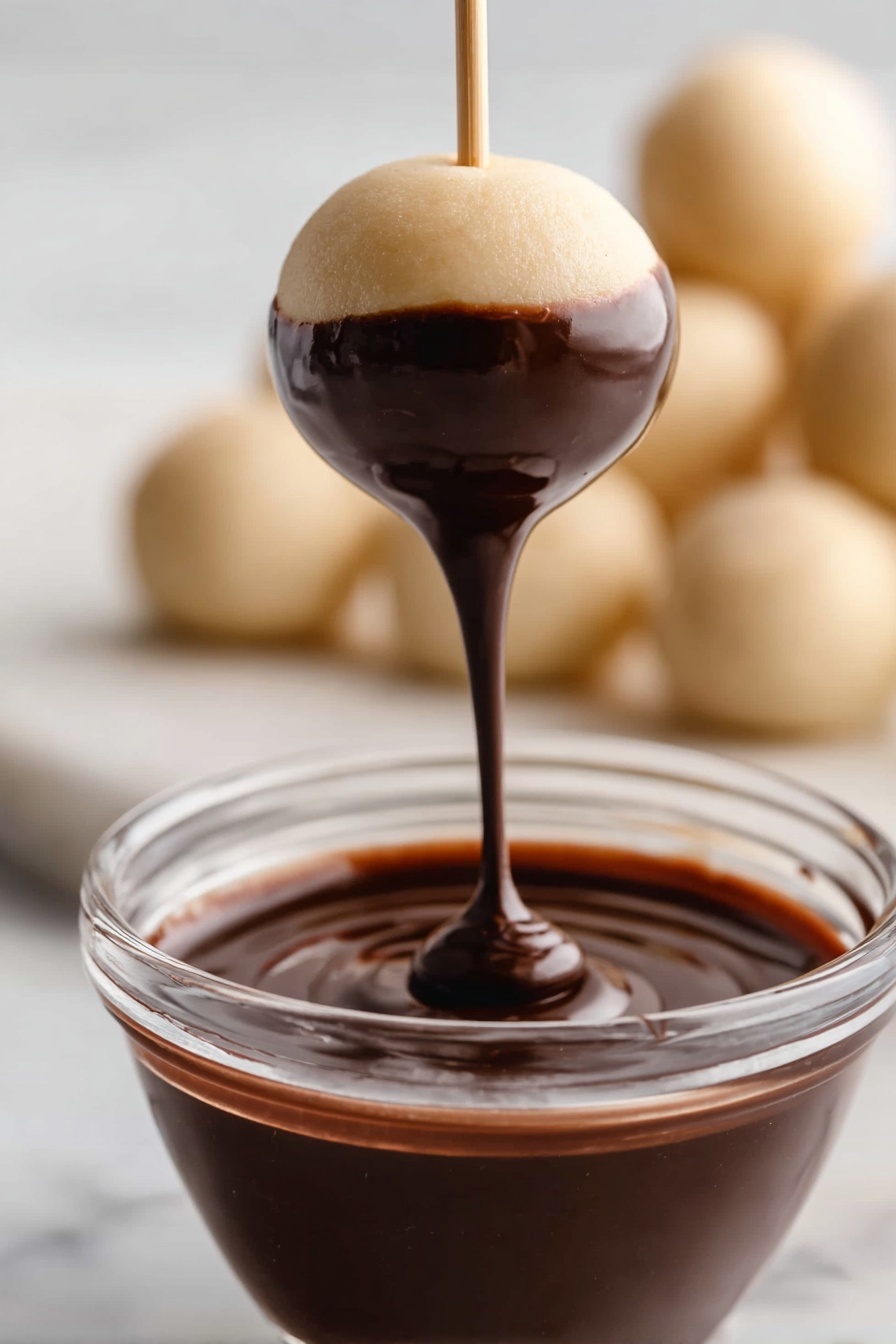 A round dough ball on a thin stick is dipped into dark, shiny chocolate sauce, covering about half of the ball with smooth, glossy chocolate. Below the ball is a clear glass bowl filled halfway with the same thick chocolate. In the blurred background, more plain dough balls are placed on a white marbled surface, giving a soft and light contrast to the rich chocolate. photo taken with an iphone --ar 2:3 --v 7 - Chocolate-Dipped Peanut Butter Balls, Buckeyes, no-bake peanut butter treats, easy chocolate peanut butter balls, simple no-bake dessert