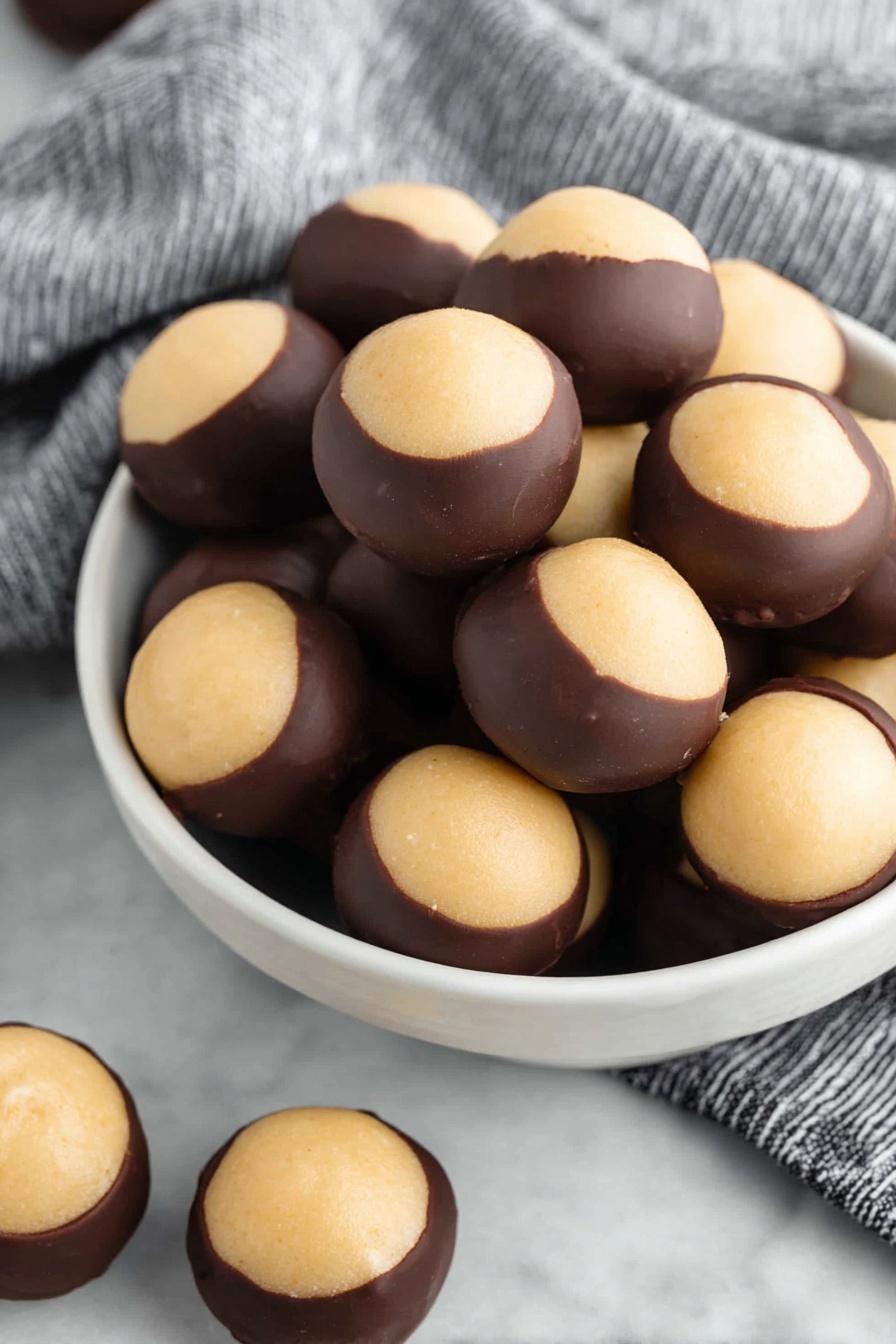 A white bowl filled with round treats that have two main layers: the bottom layer is dark brown and smooth like chocolate, covering about two-thirds of each ball, while the top layer is a light beige, creamy and slightly textured, covering the remaining one-third at the top center. The treats are clustered together in the bowl, with more of the same treats scattered on a white marbled surface around the bowl, which sits on a striped cloth. photo taken with an iphone --ar 2:3 --v 7 - Chocolate-Dipped Peanut Butter Balls, Buckeyes, no-bake peanut butter treats, easy chocolate peanut butter balls, simple no-bake dessert