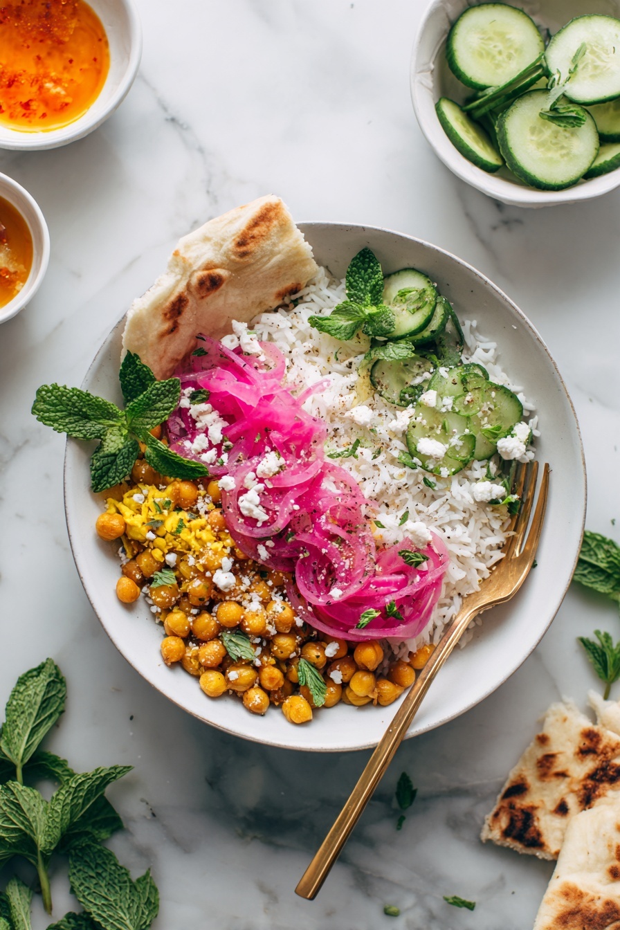 The image shows a white bowl filled with a colorful dish that has four main layers: white rice on one side, a mixture of golden-brown chickpeas and yellow lentils in the middle, and topped with bright pink pickled onions along with fresh green cucumber slices and mint leaves. There are small white crumbles of cheese scattered on top. A piece of flatbread rests along the edge of the bowl. A gold fork is placed inside the bowl on the right side. Around the bowl, there are smaller white bowls, one with cucumber slices and another with orange sauce. Pieces of flatbread and green herbs are scattered on a white marbled surface. Photo taken with an iphone --ar 2:3 --v 7 - Mediterranean Chicken Bowls, Mediterranean Chicken Bowls with Spiced Feta, easy Mediterranean chicken bowls, quick healthy dinner recipes, flavorful chicken bowls