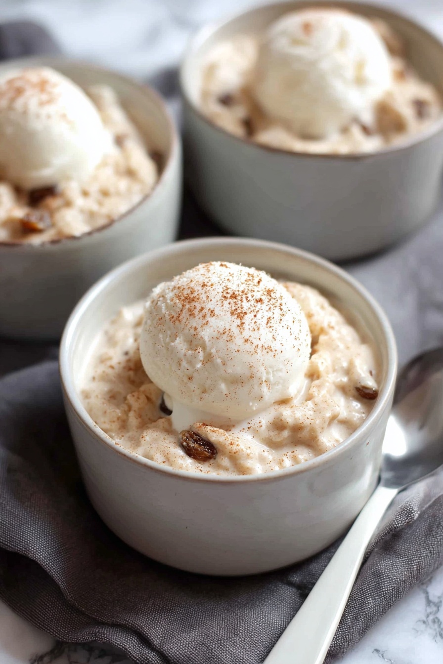 A white bowl filled with a thick creamy oatmeal mixture that has a light tan color with soft, chunky texture and dark brown raisins mixed throughout. On top of the oatmeal is a single scoop of smooth, pale white ice cream with a light dusting of brown cinnamon powder. A silver spoon is partially submerged in the oatmeal on the right side of the bowl. The bowl sits on a soft pink cloth with a white marbled surface underneath. Photo taken with an iphone --ar 2:3 --v 7 - Creamy Cinnamon Rice Pudding, easy rice pudding with cinnamon, comforting dessert recipes, homemade rice pudding, cinnamon dessert ideas