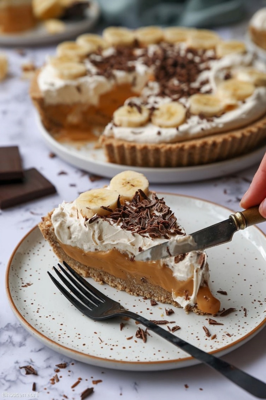 A slice of pie sits on a white plate with brown speckles, showing three layers: a crunchy light brown crust, a smooth caramel-colored filling with banana slices, and a creamy white topping sprinkled with dark chocolate curls. Behind it, the rest of the pie with the same layers is on a white plate, with one slice missing. A woman’s hand holds a silver knife with some pie filling and cream on it. A black fork rests on the plate near the slice. The background is a white marbled surface with some chocolate shavings scattered around. Photo taken with an iphone --ar 2:3 --v 7 - Easy No-Bake Banoffee Pie, no-bake banana dessert, simple banana pie, quick banoffee pie, effortless dessert recipes