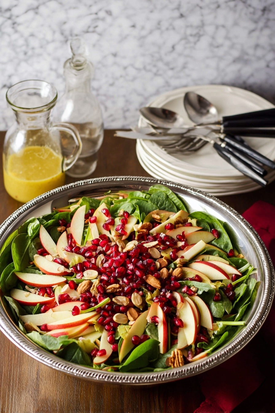 A large silver bowl holds a fresh salad with three main layers: the bottom layer is dark green spinach leaves with smooth texture, the middle layer is thin, light cream and red slices of apple arranged evenly over the spinach, and the top layer has bright red pomegranate seeds and small pieces of light tan nuts sprinkled throughout. The bowl is on a wooden surface with a white marbled texture background. Nearby are two silver serving spoons, a small glass jar of yellow dressing, and a stack of white plates with black-handled forks. photo taken with an iphone --ar 2:3 --v 7 - Apple Cranberry Spinach Salad, healthy fruit and spinach salad, easy fall salad recipe, quick festive salad, colorful nutritious salad