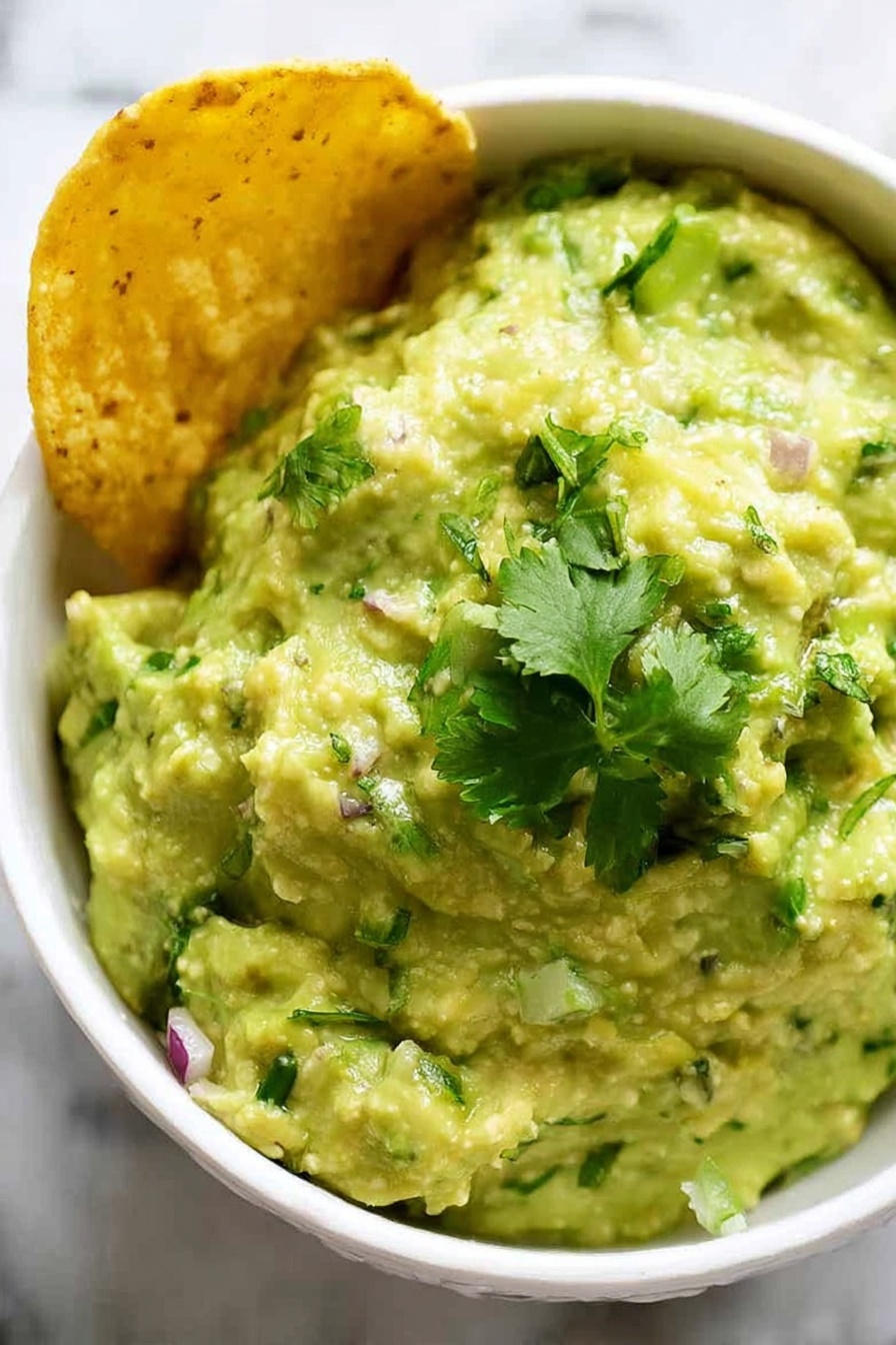 A close-up view of a white bowl filled with creamy, chunky pale green guacamole mixed with small bits of green herbs and red onion, topped with fresh bright green cilantro leaves. On the left side of the bowl, a golden yellow corn chip is partially dipped into the guacamole. The bowl sits on a white marbled surface. photo taken with an iphone --ar 2:3 --v 7 - Creamy Guacamole without Recipe, Fresh Guacamole Ingredients, Easy Guacamole Dip, Best Homemade Guacamole, Healthy Avocado Dip