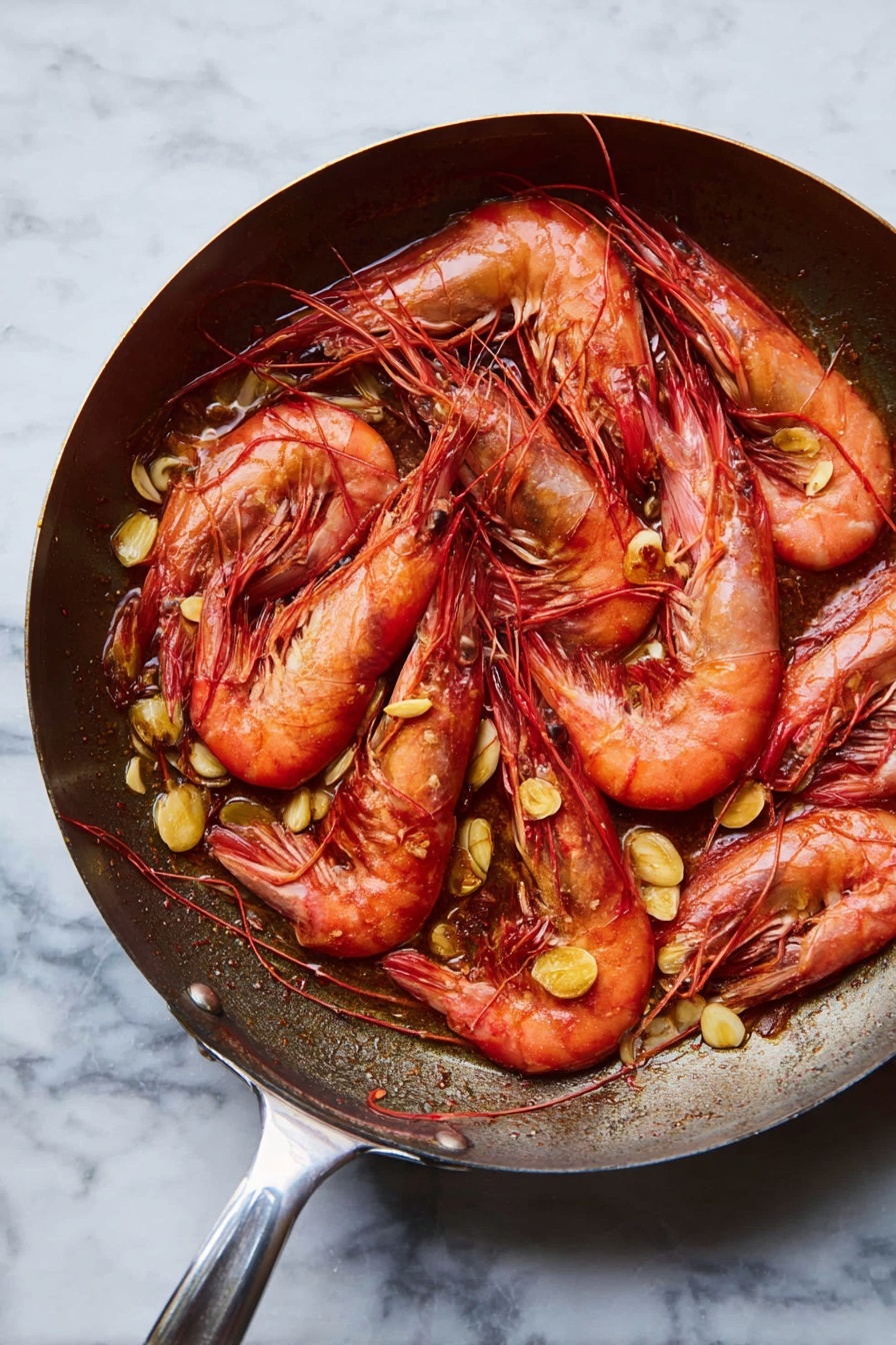 A frying pan filled with cooked whole red prawns layered over scattered golden brown slices of sautéed garlic. The prawns are arranged in a slightly overlapping way, showing their curved, shell-covered bodies, long antennae, and black eyes. The pan has a shiny metal handle extending to the side, and the background is a white marbled surface. photo taken with an iphone --ar 2:3 --v 7 - Garlic Sautéed Shrimp, Shrimp sautéed with garlic, Easy shrimp recipes, Quick seafood dishes, Spanish-inspired shrimp
