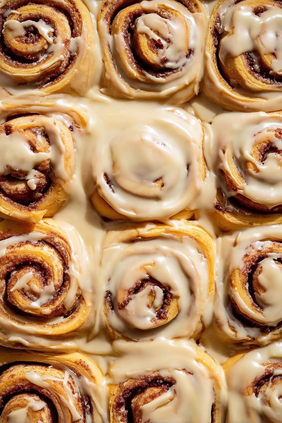 The image shows a close-up view of twelve cinnamon rolls tightly placed in a white pan, each roll having a spiral shape with layers of soft, golden-brown dough and dark cinnamon filling in between. The rolls are generously covered with smooth, creamy, light beige icing that spreads over the top and drips down slightly onto the sides and edges of each roll. The texture of the dough looks soft and fluffy, while the cinnamon filling adds a rich contrast in color. The light brown icing has a glossy finish, adding a sense of sweetness and warmth to the whole scene. The white marbled surface beneath complements the warm tones of the cinnamon rolls. photo taken with an iphone --ar 2:3 --v 7 - Gingerbread Cinnamon Rolls, holiday cinnamon rolls, spiced Christmas breakfast, soft fluffy cinnamon rolls, gingerbread dessert ideas