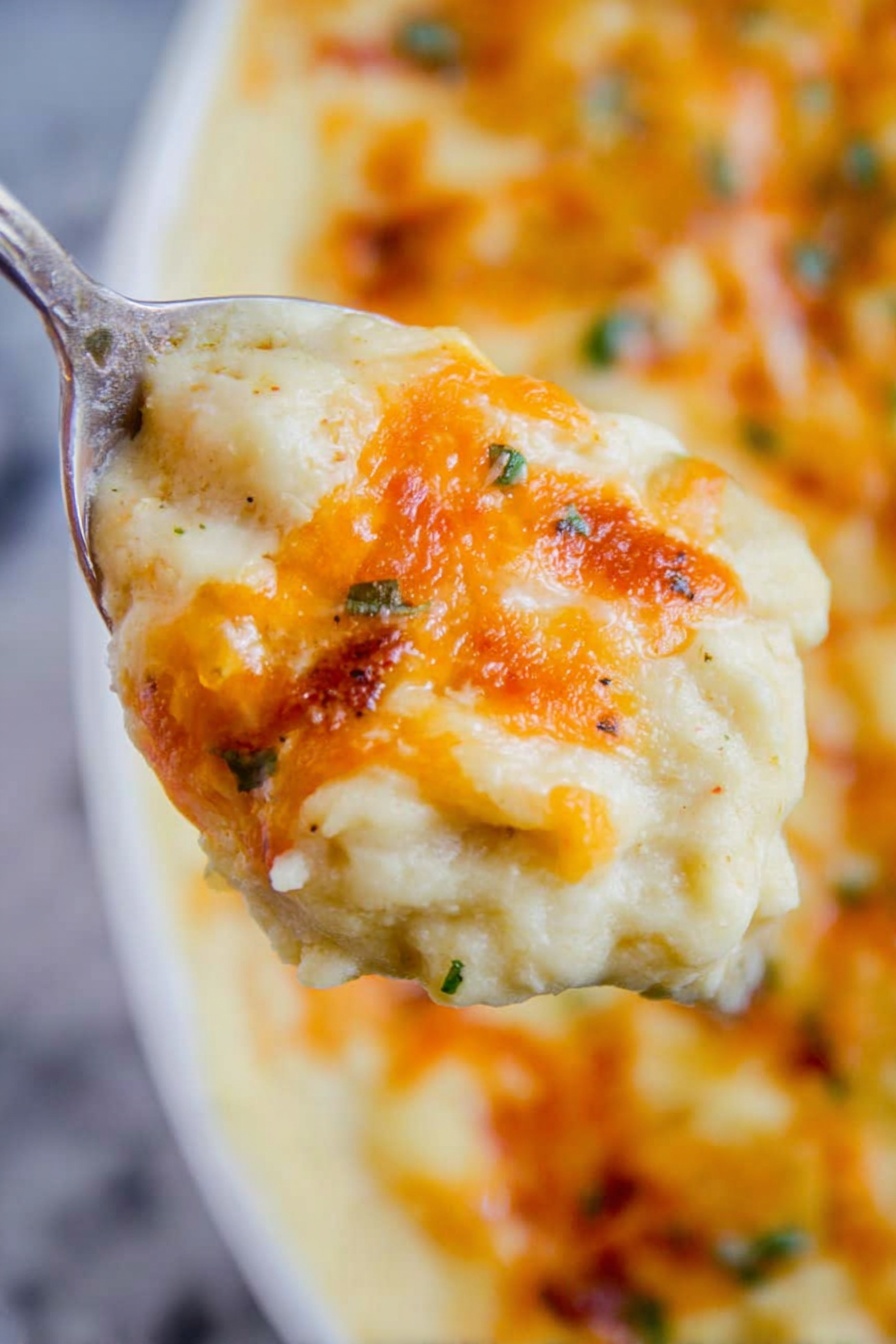 A close-up view of a spoon holding a thick, creamy dish that has a soft pale yellow base layer with a smooth texture. On top of this base, there is a layer of melted orange cheese with hints of browned spots and small green herb pieces scattered throughout. The background shows more of the same dish with a slightly uneven surface and golden brown cheese melted over it. The scene is set against a white marbled texture. Photo taken with an iphone --ar 2:3 --v 7 - Creamy Make-Ahead Mashed Potatoes, make-ahead mashed potatoes, holiday mashed potatoes, easy mashed potato recipe, cheesy mashed potatoes