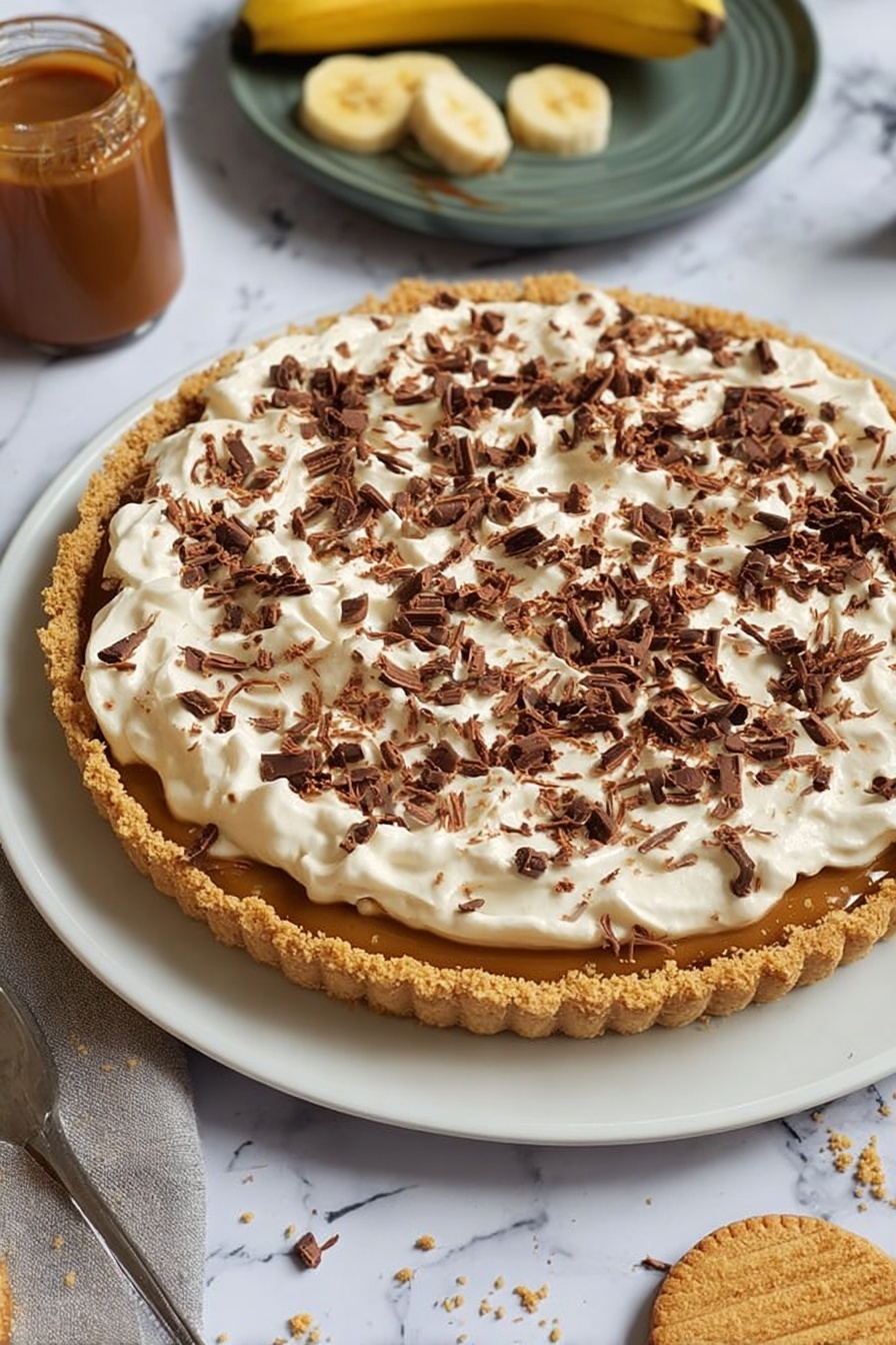 The image shows a dessert tart on a white plate with a crumbly golden brown crust forming the base, filled with a caramel-colored layer visible under a thick top layer of fluffy, white cream spread unevenly. The cream is generously covered with small, dark brown chocolate curls scattered across the surface. Around the plate, there is a jar of caramel sauce, a partially peeled banana, and a round biscuit with a bite taken out, all set on a white marbled surface. Photo taken with an iphone --ar 2:3 --v 7 - Easy No-Bake Banoffee Pie, no-bake banana dessert, simple banana pie, quick banoffee pie, effortless dessert recipes