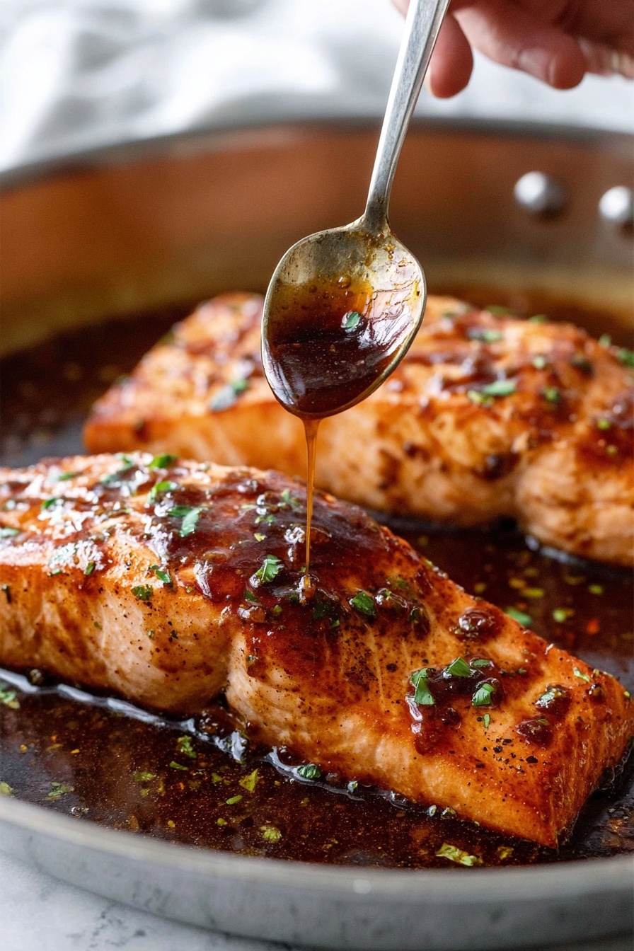Two thick salmon fillets with a shiny dark brown sauce cover. The top layer is glossy, sticky sauce with small green herb bits sprinkled over. The salmon’s surface is textured with light char marks and a golden brown color under the sauce. A woman's hand holds a metal spoon above the front fillet, drizzling more sauce from the spoon onto the fish. The fillets sit in a shallow pool of the dark sauce within a metal pan. The background has a soft, white marbled texture. photo taken with an iphone --ar 2:3 --v 7 - Honey Garlic Glazed Salmon, Honey Garlic Salmon, Easy Salmon Recipes, Healthy Salmon Dinner, Quick Seafood Recipes