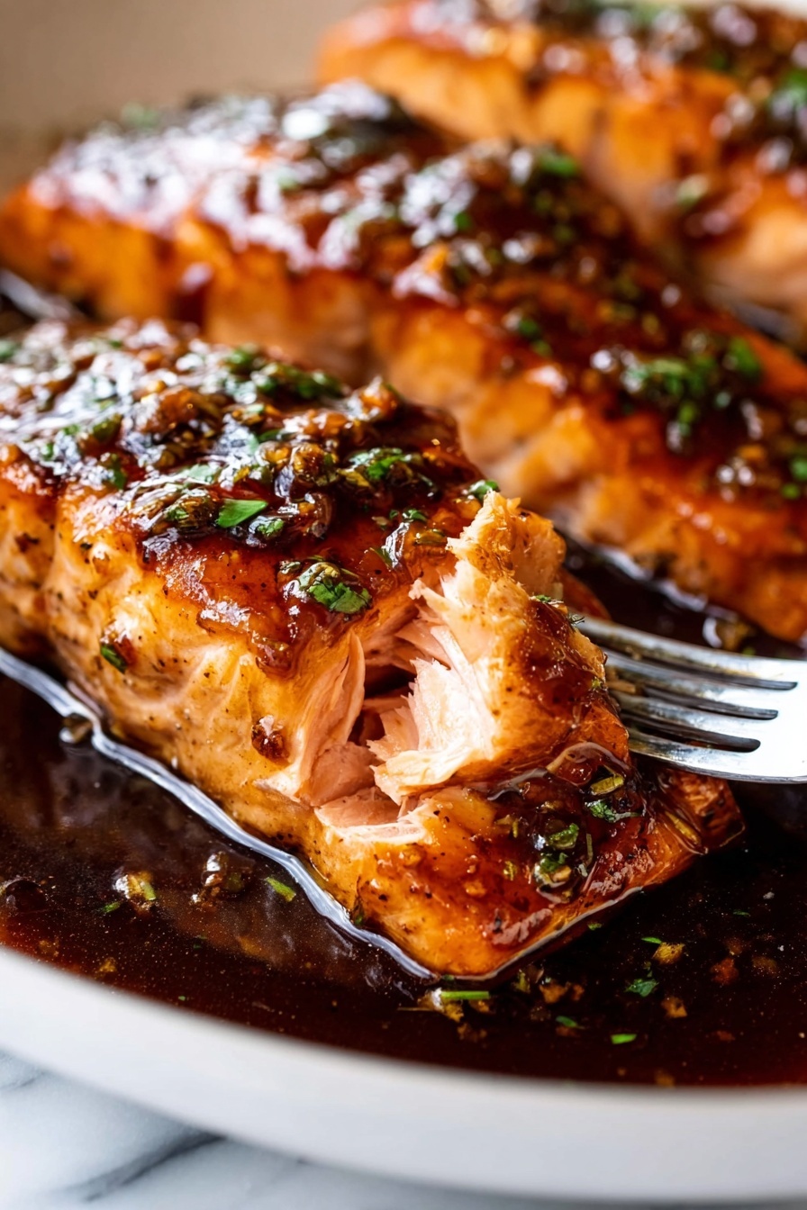 The image shows a close-up of two thick, cooked salmon pieces in a white pan, with the focus on the front piece where a fork pulls a small section showing its light pink, flaky inside layers. The salmon is covered in a shiny, dark brown sauce with a slight glaze and small green herb bits sprinkled on top. The sauce pools around the base of the salmon, reflecting light and adding a rich texture. The pan sits on a white marbled surface, and the salmon skin underneath is visible with a slightly darker color. Photo taken with an iphone --ar 2:3 --v 7 - Honey Garlic Glazed Salmon, Honey Garlic Salmon, Easy Salmon Recipes, Healthy Salmon Dinner, Quick Seafood Recipes