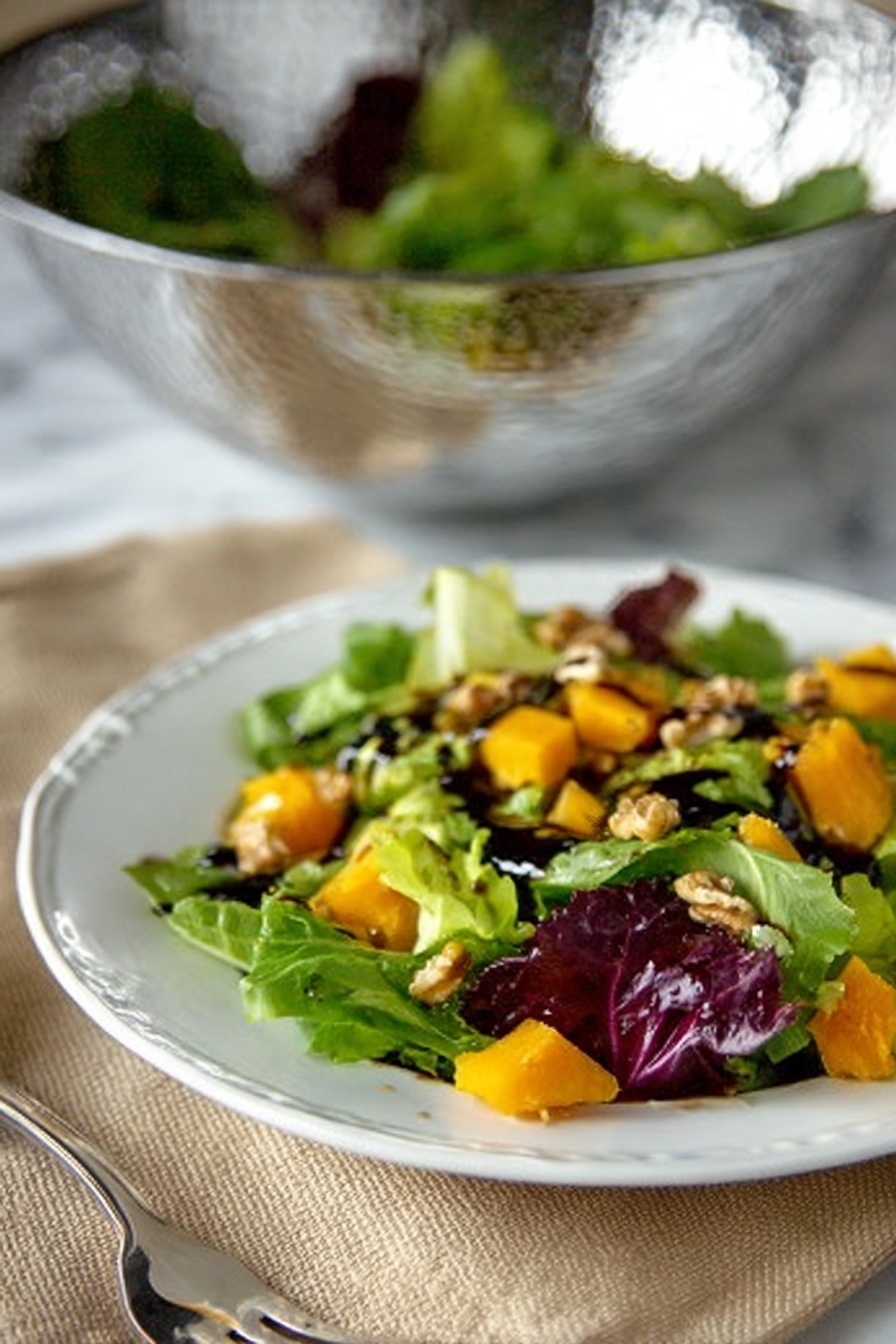 The image shows a white plate with a fresh salad on top placed on a beige cloth. The salad has several layers: a base layer of mixed green lettuce leaves that are bright green and purple with a soft texture, scattered orange chunks of roasted pumpkin or squash spread evenly over the greens, and small pieces of walnuts sprinkled throughout. Some dark balsamic dressing is drizzled over the salad, adding a glossy shine. In the background, a silver textured bowl with more greens inside sits on a white marbled surface, slightly blurred. A fork is visible next to the plate on the left side. photo taken with an iphone --ar 2:3 --v 7 - Roasted Butternut Squash Salad with Cranberries, autumn salad recipes, healthy fall salads, festive holiday side dishes, easy roasted vegetable salads