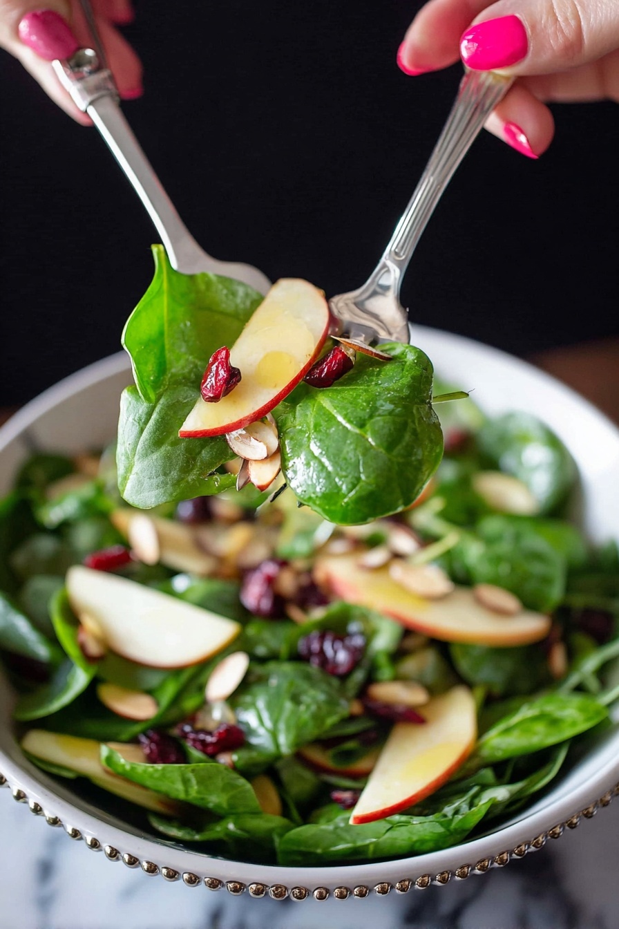 A close-up of a fresh salad being lifted by two silver utensils held by a woman's hand with pink nail polish. The salad has bright green spinach leaves, thin light yellow apple slices with red edges, small dark red dried cranberries, and light brown sliced almonds mixed inside a white bowl with a beaded rim. The background is dark, contrasting with the vibrant colors of the salad, and the surface underneath the bowl is a white marbled texture. photo taken with an iphone --ar 2:3 --v 7 - Apple Cranberry Spinach Salad, healthy fruit and spinach salad, easy fall salad recipe, quick festive salad, colorful nutritious salad