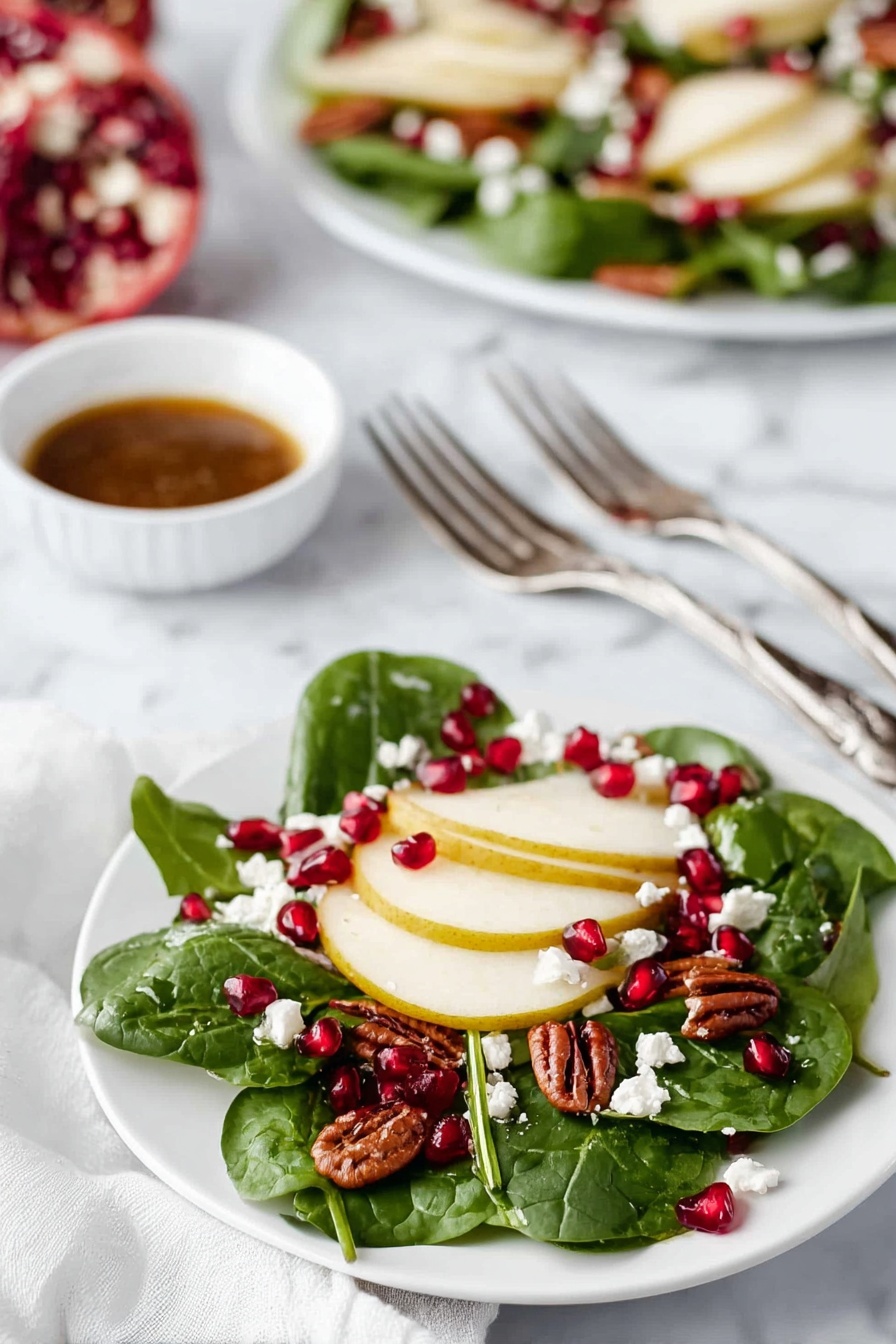 The dish shows a small white plate with a fresh salad made of three main layers. The first bottom layer consists of fresh dark green spinach leaves spread across the plate. On top of this, the second layer has thin round slices of light yellow pear arranged evenly. The third layer is made up of small bright red pomegranate seeds scattered over the pears and spinach, along with small white crumbly pieces of cheese, and a few medium brown pecan nuts. In the background, there is a larger white plate with the same salad and a small white bowl filled with a brown dressing in the center, all set on a white marbled surface. Near the small plate are two silver forks resting on a white cloth. photo taken with an iphone --ar 2:3 --v 7 - Christmas Wreath Spinach Salad with Pomegranate, festive holiday salad, healthy Christmas salad, easy holiday spinach salad, seasonal fruit salad