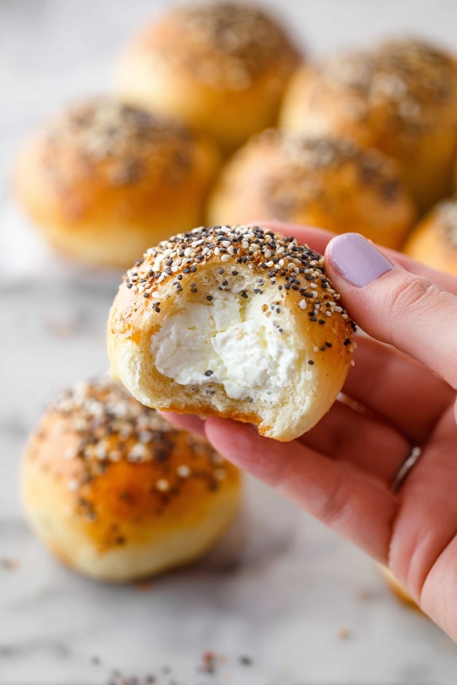 A close-up view of a woman's hand holding a small round bread roll with a golden brown crust topped with a mix of black and white seeds. The roll has a bite taken out, revealing a soft white inside filled with creamy white cheese. In the background, several similar bread rolls are placed on a white marbled surface, each with the same seed topping, slightly out of focus. Photo taken with an iphone --ar 2:3 --v 7 - Stuffed Bagel Bites with Cream Cheese, Bagel Bites recipe, Easy bagel snacks, Cream cheese stuffed bagels, Quick appetizer ideas
