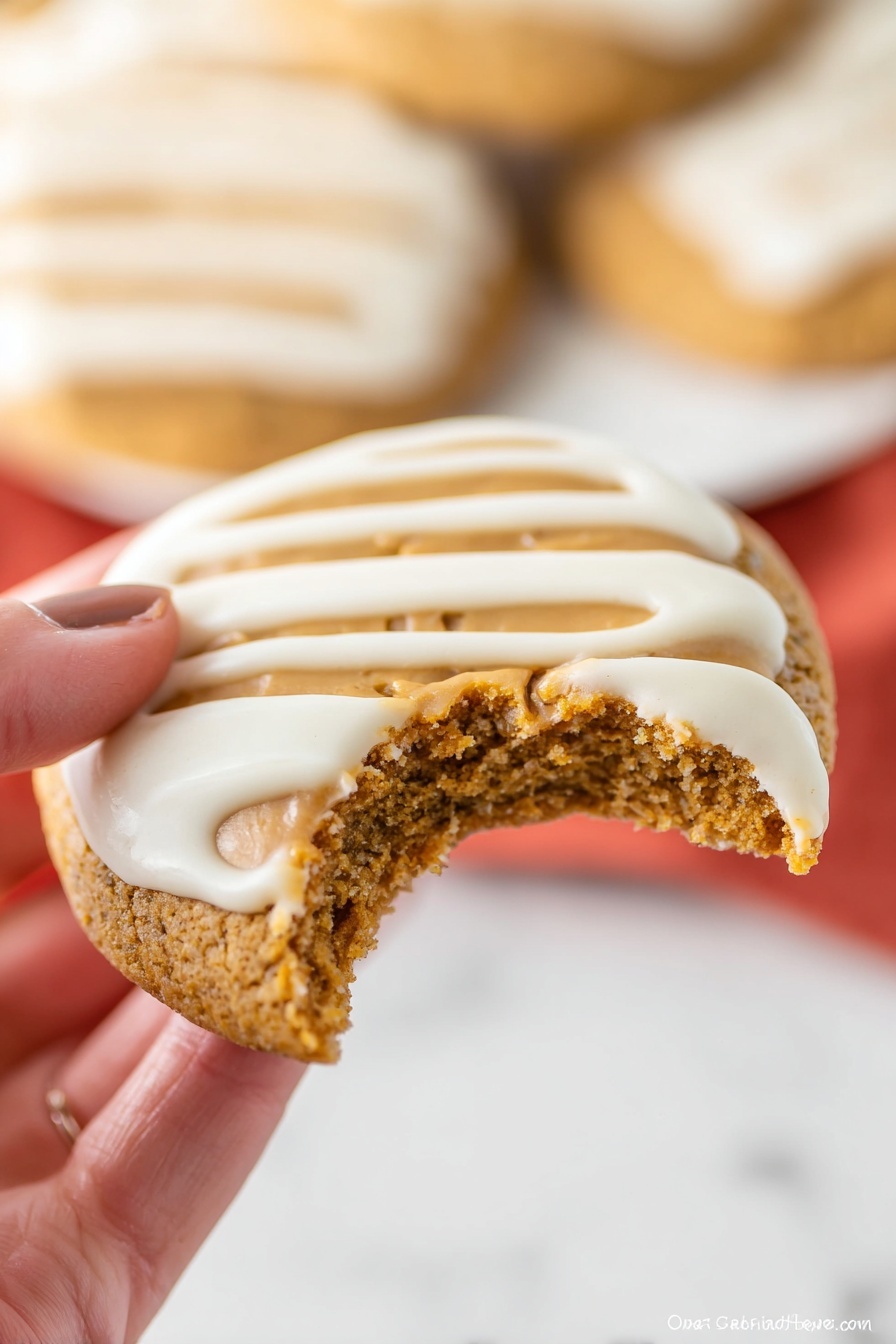 A close-up of a round cookie held by a woman's hand, showing a bite taken from the bottom right edge. The cookie has a soft, slightly crumbly golden brown base with a smooth, light brown peanut butter layer spread in the center. On top, three thick white icing stripes run horizontally across the cookie's surface. The background shows another cookie with similar white icing on a white marbled surface but blurred. Photo taken with an iphone --ar 2:3 --v 7 - Gingerbread Thumbprint Cookies with White Chocolate, holiday gingerbread cookies, white chocolate filled cookies, festive thumbprint cookies, easy holiday baking recipes