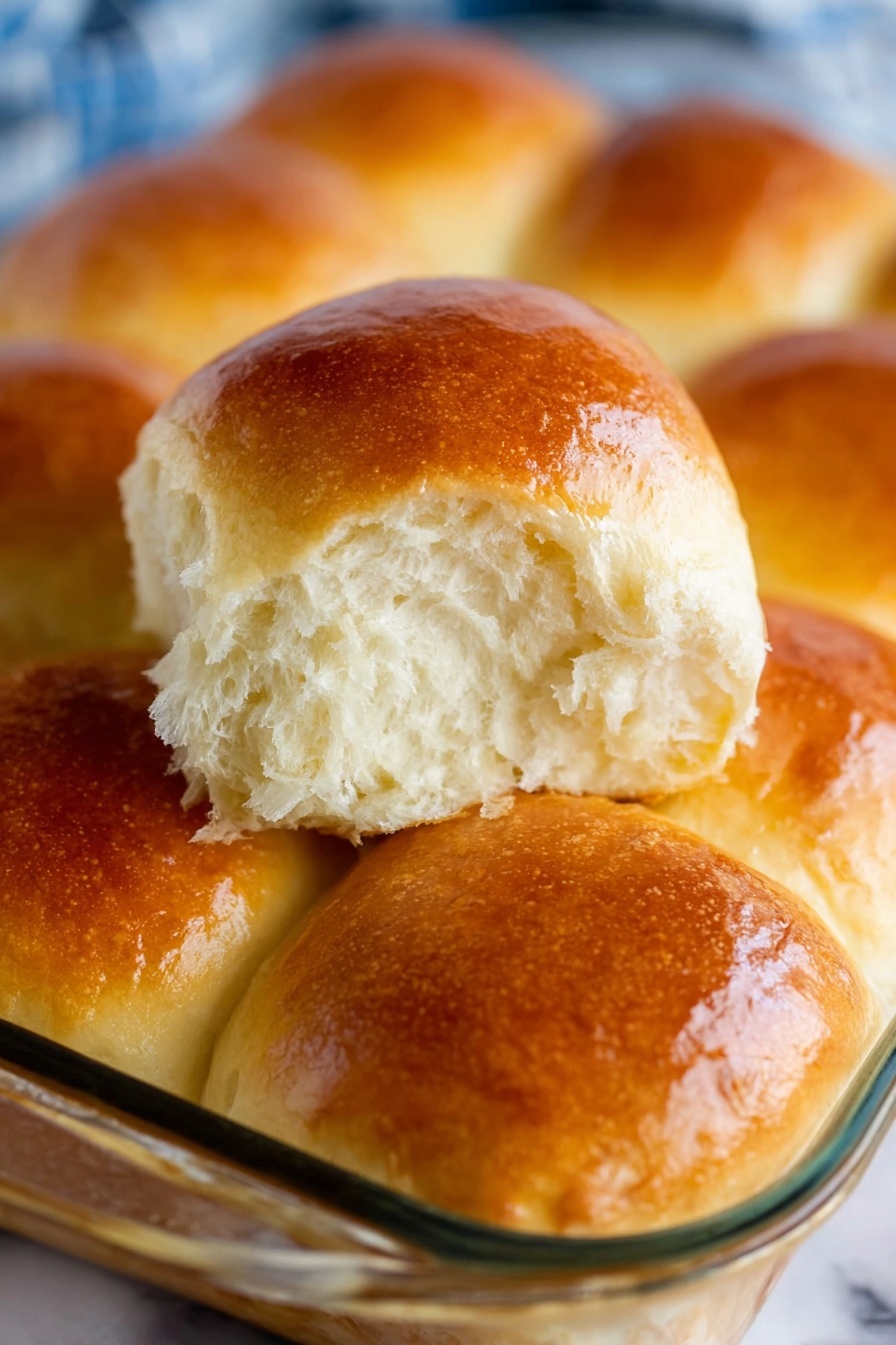 The image shows a group of soft, shiny, golden-brown dinner rolls placed close together in a clear glass baking dish. The rolls are bread-colored with a smooth, slightly glossy top surface that reflects light, showing a fresh and fluffy texture inside. One roll is lifted up, revealing a white, airy, and light inside, contrasting with the warm brown crust. The background is a white marbled surface, and the focus is on the detailed texture of the bread rolls. photo taken with an iphone --ar 2:3 --v 7 - Fluffy Potato Rolls, soft bread rolls, homemade potato rolls, easy bread recipes, tender dinner rolls