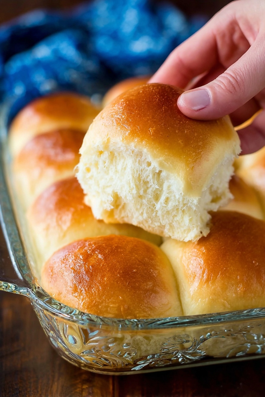 A close-up image of a woman's hand lifting one golden brown dinner roll from a glass baking dish filled with soft, fluffy rolls. The rolls have a shiny, smooth top with a light brown color, and the inside shows a soft, airy texture with a pale cream color. The glass dish has decorative patterns and is placed on a dark wooden surface with a blurred blue cloth in the background. Photo taken with an iphone --ar 2:3 --v 7 - Fluffy Potato Rolls, soft bread rolls, homemade potato rolls, easy bread recipes, tender dinner rolls
