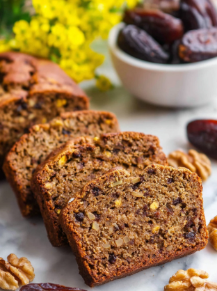 A close-up of four slices of brown nut bread lined up diagonally on a white marbled surface. Each slice shows a dense texture with visible dark brown nuts and darker bits inside the bread. Around the slices are loose whole walnuts and dark brown dried dates scattered. In the background, there is a white bowl filled with more dried dates and a soft-focused cluster of yellow flowers. The scene is bright and natural. photo taken with an iphone --ar 2:3 --v 7 - Date Nut Bread, moist date bread with walnuts, easy homemade date nut bread, cozy loaf recipe, sweet breakfast bread with dates