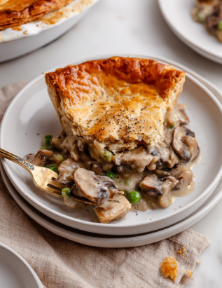 A white round plate holds a portion of vegan mushroom pot pie with two clear layers: the bottom layer is a creamy mushroom filling with sliced brown mushrooms, green peas, and a thick sauce, while the top layer is a golden-brown flaky crust slightly cracked with a sprinkled touch of black pepper. A bronze fork on the plate lifts a piece of the mushroom filling, showing its chunky texture. The plate sits on a beige cloth with a small part of a white marbled surface visible around it. In the background, part of a large round pot pie with a well-baked golden crust is visible on the left, and a white plate with another piece of pie shows on the right. Photo taken with an iphone --ar 2:3 --v 7 - Vegan Mushroom Pot Pie, vegan mushroom pie, plant-based mushroom pot pie, dairy-free mushroom pot pie, vegetarian mushroom casserole