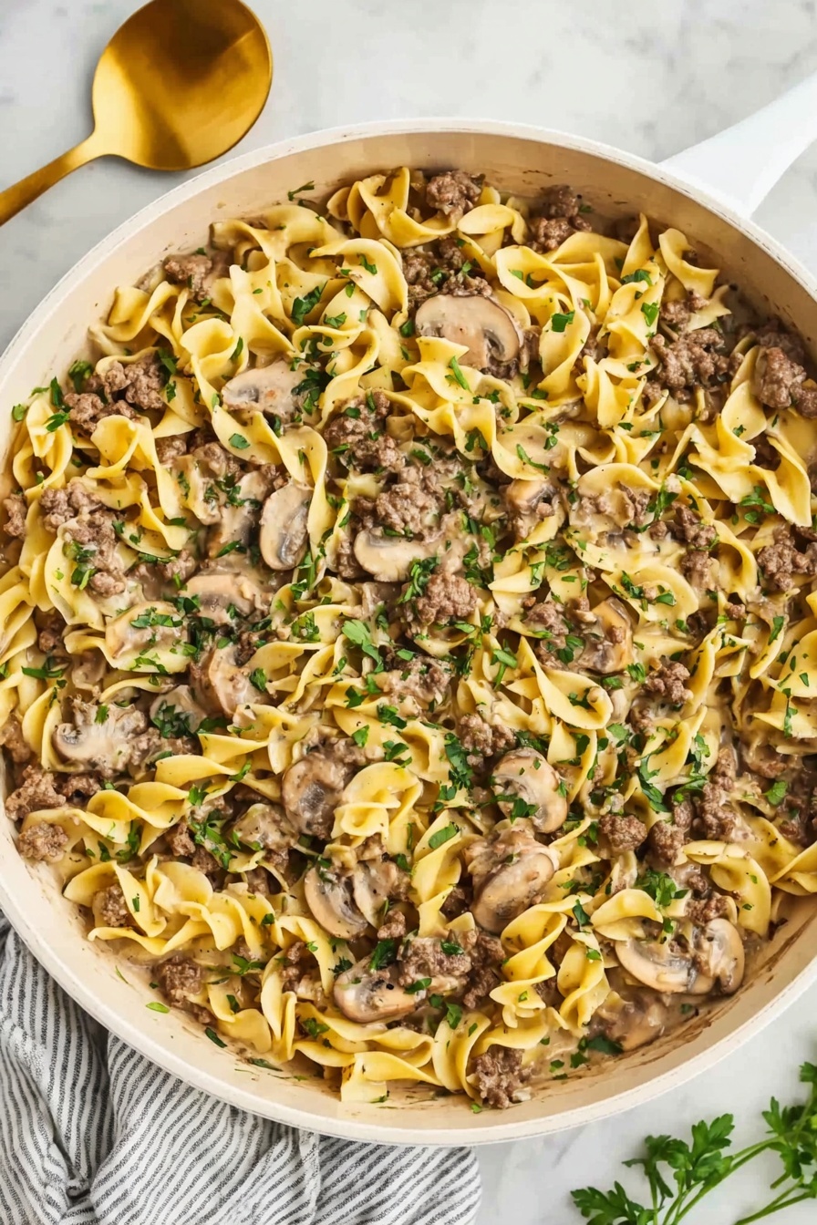 The image shows a wide white pan filled with creamy egg noodles mixed with sliced mushrooms and small pieces of cooked ground beef. The noodles are soft yellow with a smooth texture, spread evenly across the pan. The mushrooms, pale brown and slightly shiny, are mixed throughout the noodles. The beef is browned and scattered on top and inside the noodles in small chunks. Fresh green herbs are sprinkled over the whole dish, adding a pop of color. The pan sits on a white marble surface, next to a gold spoon and white cloth with gray stripes. Photo taken with an iphone --ar 2:3 --v 7 - One-Pot Ground Beef Stroganoff, ground beef stroganoff recipe, easy beef stroganoff dinner, creamy beef skillet meal, weeknight beef pasta