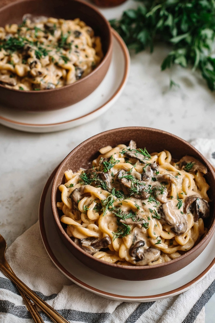 Two bowls of creamy pasta with mushroom pieces and green herbs on top are shown. Each bowl is brown and sits on a white plate with a thin brown rim, placed on a white cloth with gray stripes. The pasta is light yellow and swirled, covered in a smooth creamy sauce mixed with darker brown mushrooms and sprinkled with chopped green herbs for freshness. The setup is on a white marbled surface with some blurred green leaves in the background, giving a cozy and natural vibe. Photo taken with an iphone --ar 2:3 --v 7 - Creamy Vegan Mushroom Stroganoff, vegan mushroom pasta, dairy-free mushroom sauce, plant-based comfort food, vegan mushroom dinner