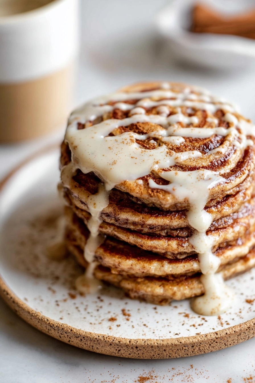 A stack of six thick pancakes sits on a white plate with brown speckles, each pancake showing swirled layers of cinnamon brown and light beige batter. The top pancake is covered with creamy white icing drizzled unevenly, some dripping down the sides and pooling on the plate. The stack rests on a white marbled surface with some loose brown cinnamon powder scattered nearby. In the blurred background, a white cup with a tan base is faintly visible. photo taken with an iphone --ar 2:3 --v 7 - Vegan Cinnamon Swirl Pancakes, gluten-free vegan breakfast, plant-based cinnamon pancakes, healthy cinnamon pancake recipe, vegan breakfast ideas