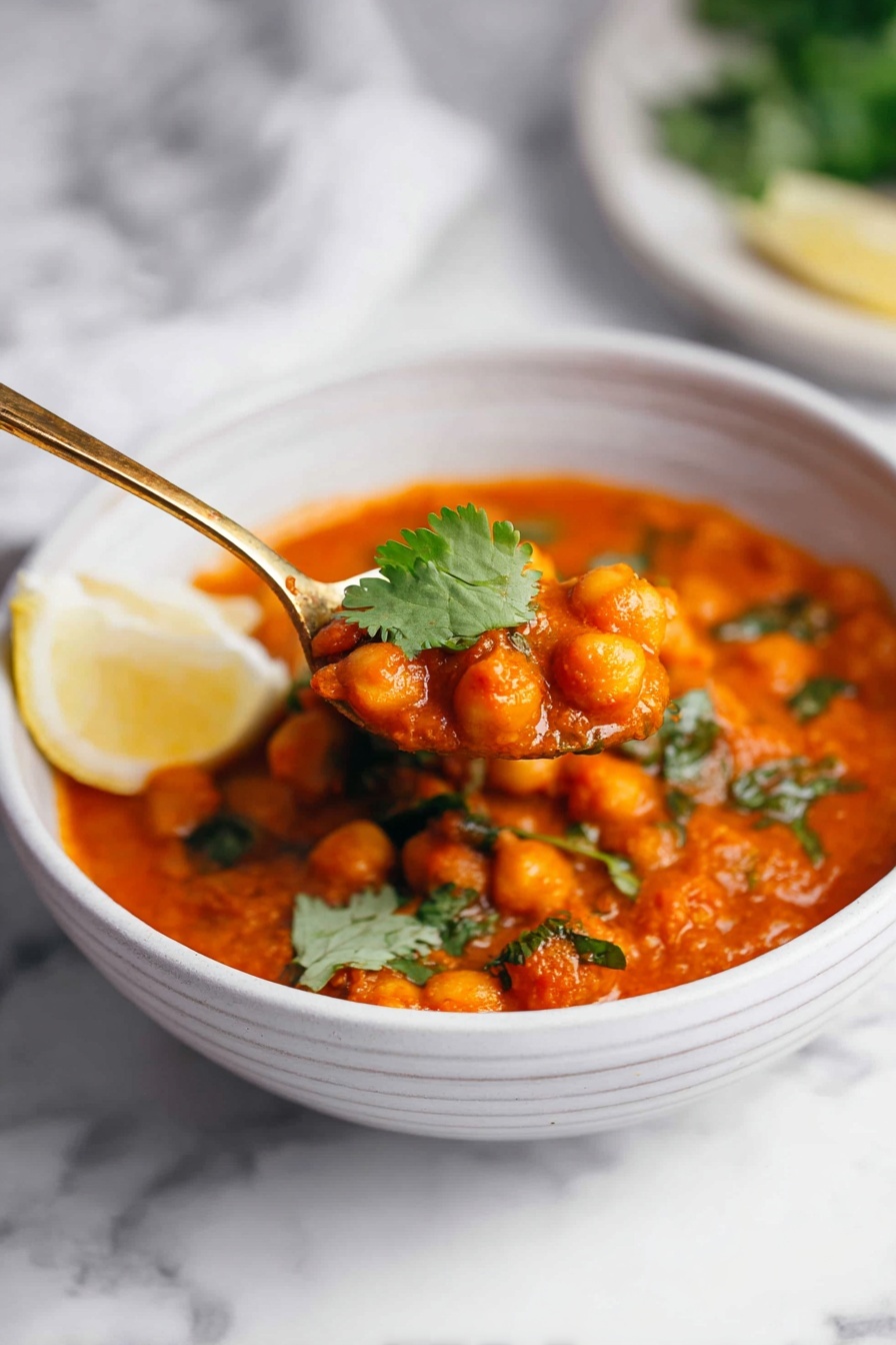 A white bowl on a white marbled surface holds a thick, orange-red chickpea curry with a slightly chunky texture. The curry is topped with fresh green cilantro leaves and a wedge of pale yellow lemon rests on the side inside the bowl. A golden spoon lifts a portion of the curry, showing the round, soft chickpeas covered in the rich sauce, with a small cilantro leaf on top. In the blurred background, a white plate with more greens and lemon wedges is visible. Photo taken with an iphone --ar 2:3 --v 7 - Creamy Coconut Chickpea Curry, vegan chickpea curry, easy coconut curry, veggie curry with coconut, one-pot chickpea curry