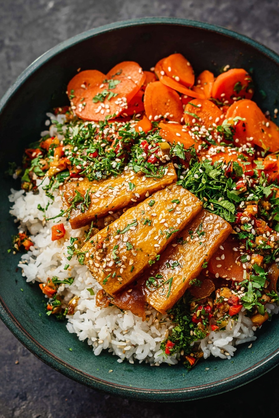 The image shows a dark green bowl filled with three main layers. The bottom layer is a bed of white rice with a soft texture. On top of the rice, there are several golden-brown rectangular pieces that look crispy on the edges, layered evenly. The top layer consists of thin orange carrot slices, small bits of green herbs, and diced vegetables mixed with sesame seeds, scattered evenly over the bowl. The whole dish looks fresh with vibrant colors and textures. Photo taken with an iphone --ar 2:3 --v 7 - Flavorful Tofu Stir Fry, tofu stir fry recipe, healthy stir fry dish, crispy tofu with savory sauce, vegan tofu stir fry