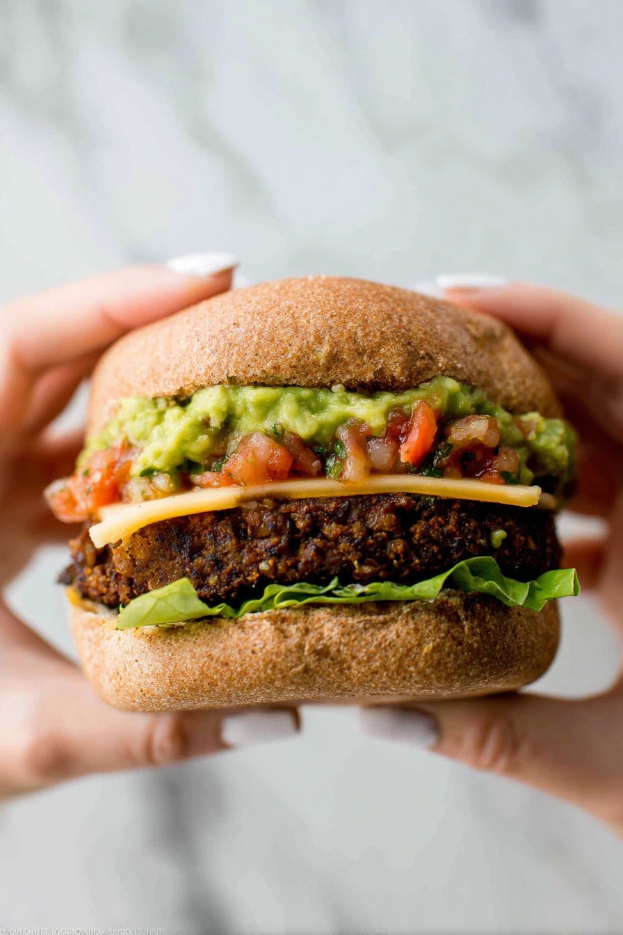 A close-up image of a sandwich held by two woman's hands with light-colored nail polish, showing four layers inside a soft, brown whole wheat bun. The bottom layer is green leaf lettuce, followed by a thick, dark brown veggie patty with a coarse, chunky texture. On top of the patty is a slice of pale yellow cheese partially melted, then a layer of chunky green guacamole with a creamy texture partly mixed with bright red tomato salsa. The top bun is slightly squished down over the layers. The background features a white marbled texture. photo taken with an iphone --ar 2:3 --v 7 - Best Black Bean Burgers, black bean burger recipe, healthy veggie burgers, vegetarian burger ideas, smoky spicy bean patties