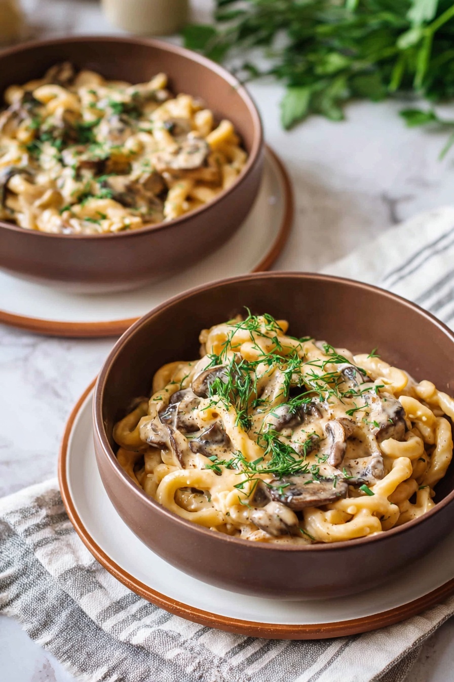 In the image, a brown bowl holds creamy pasta with ruffled edges, mixed with sliced mushrooms and garnished with small green herbs scattered on top. The bowl sits on a white plate with a thin brown rim, accompanied by a silver fork on a light-colored cloth under the plate. The background shows a white marbled texture with another similar bowl slightly blurred in the back, creating a cozy and warm feeling. photo taken with an iphone --ar 2:3 --v 7 - Creamy Vegan Mushroom Stroganoff, vegan mushroom pasta, dairy-free mushroom sauce, plant-based comfort food, vegan mushroom dinner