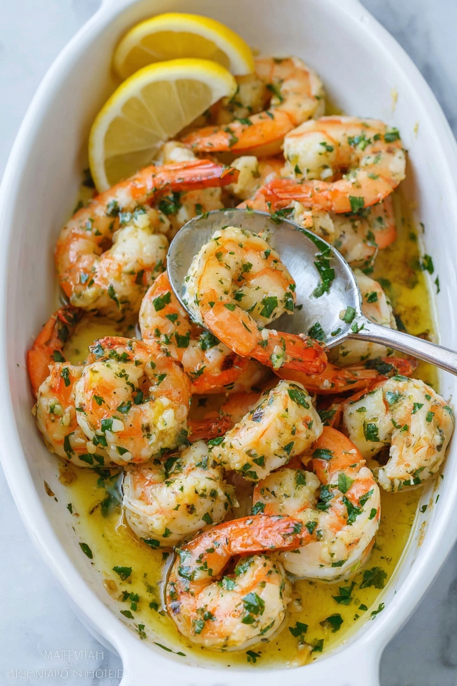 In a white oval dish filled with plump cooked shrimp, the shrimp are arranged in a loose pile with orange-pink tails curled and white meaty bodies showing. They are coated in a glossy yellow garlic butter sauce mixed with bright green chopped herbs scattered on top and mixed throughout. A silver spoon inside the dish holds several shrimp, partially covered in sauce, as two lemon wedges sit beside the dish on a white marbled surface. The shrimp have a lightly browned texture and there are small bits of garlic visible on them. Photo taken with an iphone --ar 2:3 --v 7 - Easy Shrimp Scampi with Garlic and White Wine, shrimp scampi, garlic shrimp recipe, quick seafood dinner, white wine shrimp pasta