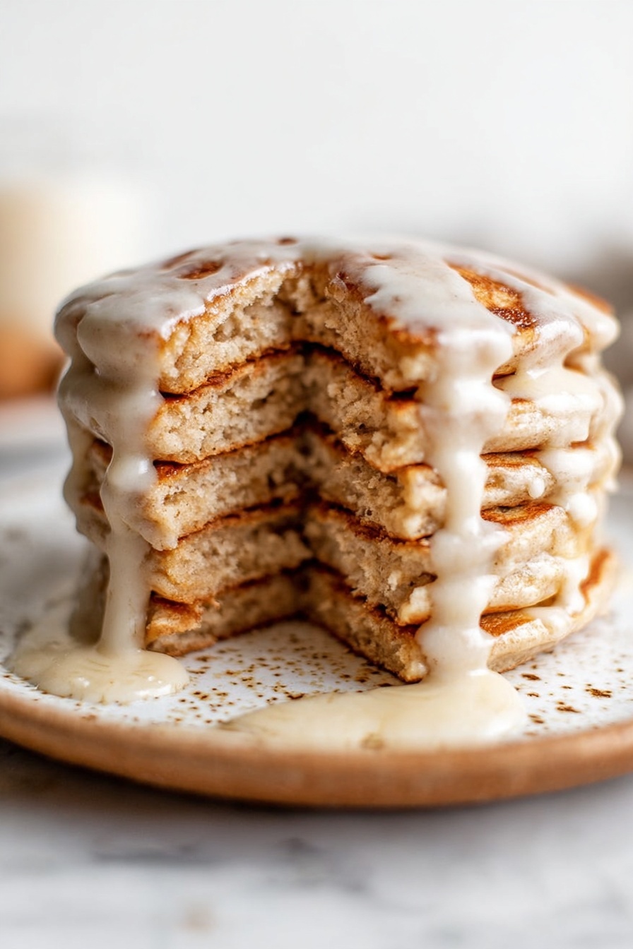 A stack of five thick pancakes with a light brown color and a slightly rough texture is placed on a white plate with brown specks and smudges. Creamy white glaze is poured over the top, dripping down unevenly between the layers and pooling slightly at the bottom. The pancakes show a soft and fluffy inside with visible air pockets. The plate is set on a white marbled surface. Photo taken with an iphone --ar 2:3 --v 7 - Vegan Cinnamon Swirl Pancakes, gluten-free vegan breakfast, plant-based cinnamon pancakes, healthy cinnamon pancake recipe, vegan breakfast ideas