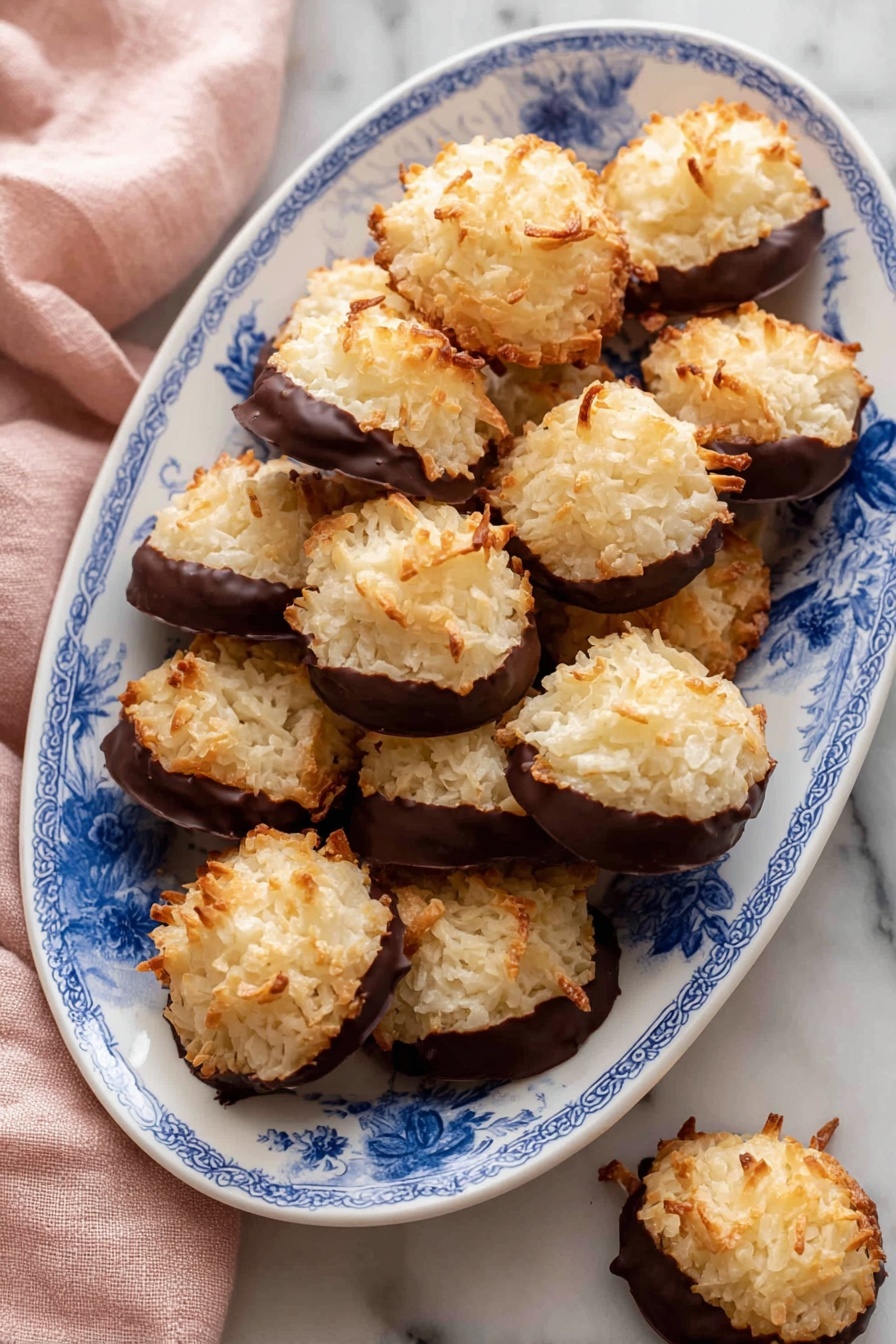 The image shows a white oval plate with a blue floral pattern filled with round coconut cookies. Each cookie has two layers: the top layer is golden brown with a rough, shredded texture of toasted coconut, and the bottom layer is dipped in smooth, dark chocolate that covers about half the cookie. The cookies are piled on the plate over a white marbled surface, with a soft pink cloth partially visible on the left side. One cookie is set just outside the plate at the bottom right. The photo taken with an iphone --ar 2:3 --v 7 - Chocolate-Dipped Coconut Macaroons, coconut macaroons recipe, easy coconut cookies, chocolate-coated macaroons, chewy coconut treats