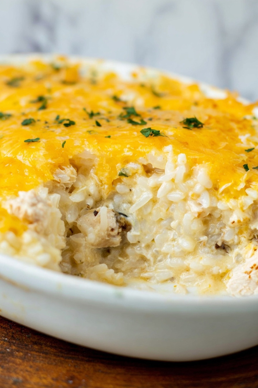 The dish shows a close-up of a creamy cheesy casserole in a white dish, featuring visible layers where the top layer is melted golden-yellow cheese with some small green herb bits sprinkled on it. Below the cheese, there is a thick layer of creamy rice mixed with white chunks of cooked meat and bits of darker ingredients, giving a soft texture with a mix of white, light yellow, and brown shades. The white dish rests on a wooden surface, but the background is changed to a white marbled texture. photo taken with an iphone --ar 2:3 --v 7 - Creamy Chicken and Rice Casserole, easy comfort food recipes, hearty baked chicken and rice dish, quick dinner ideas, cheesy casserole recipes