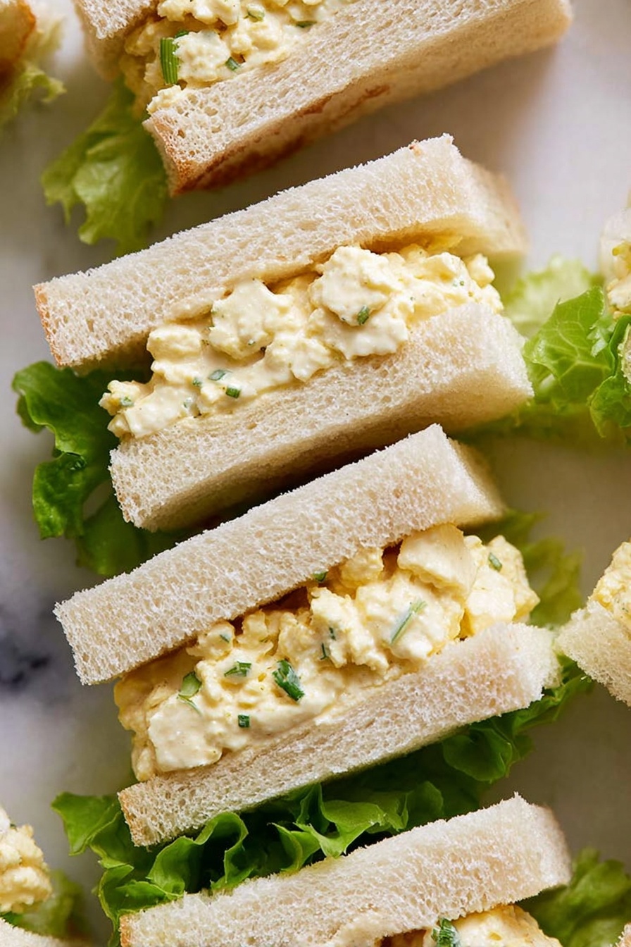 The image shows small sandwich pieces with two layers of soft, white bread surrounding a creamy, pale yellow egg salad filling mixed with small bits of egg white and sprinkled with green herbs. The sandwiches are placed on top of bright green lettuce leaves, which add a fresh contrast to the light-colored sandwiches. The texture of the bread looks soft and slightly fluffy, while the filling appears smooth but textured with tiny egg pieces. The background surface is a white marbled texture. photo taken with an iphone --ar 2:3 --v 7 - Creamy Soft Boiled Egg Sandwich,soft boiled egg breakfast ideas,easy egg sandwich recipe,creamy eggs on bread,quick egg sandwich for breakfast