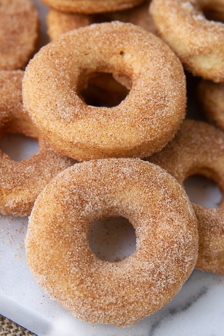 A close-up view of several round donuts with a light golden color, covered evenly with a fine layer of cinnamon sugar. The donuts have a soft texture with a slightly rough surface due to the sugar coating. They are resting directly on a shiny white marbled surface, with some donuts overlapping each other, creating a cozy piled look. The photo is naturally lit, showing subtle shadows that enhance the round shape and sugar texture of the donuts, photo taken with an iphone --ar 2:3 --v 7 - Baked Cinnamon Doughnuts, cinnamon doughnuts, baked doughnut recipe, easy baked doughnuts, cinnamon sugar doughnuts