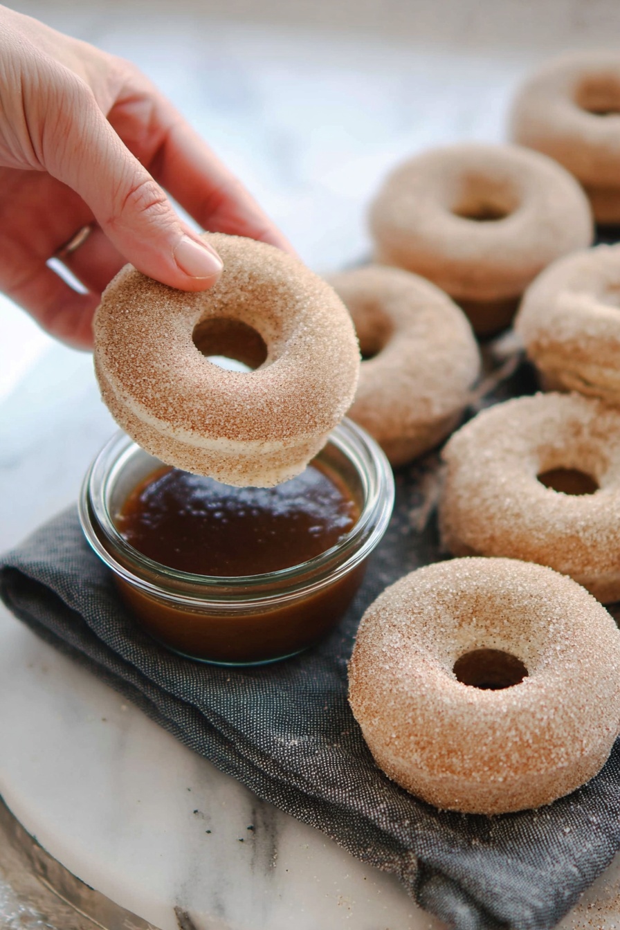 A white marbled surface holds a dark gray cloth with seven round donuts covered in light brown sugar, each with a smooth, grainy texture. At the front, a woman's hand gently holds one donut above a small, clear glass jar filled with a glossy, deep brown liquid. The donut has a round hole in the middle and is slightly lifted as if about to be dipped. The background has a soft, bright ambiance. photo taken with an iphone --ar 2:3 --v 7 - Baked Cinnamon Doughnuts, cinnamon doughnuts, baked doughnut recipe, easy baked doughnuts, cinnamon sugar doughnuts