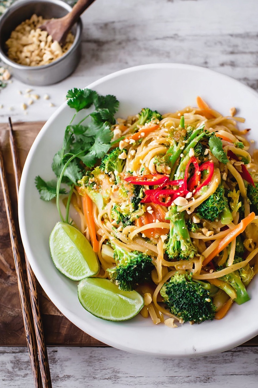 A white plate holds a colorful dish of stir-fried noodles layered with bright green broccoli florets and thin orange carrot strips, mixed with light yellow noodles and thin slices of red chili peppers scattered on top. The noodles are garnished with fresh green sprouts and crushed peanuts. On one side of the plate, two lime wedges add a fresh, bright yellow-green color. A few cilantro leaves sit on top, adding deep green contrast. The plate rests on a white marbled surface, with chopsticks placed near the back side. In the background, there is a small metal bowl with crushed peanuts and a wooden spoon. Photo taken with an iphone --ar 2:3 --v 7 - Vegan Pad Thai, vegan Pad Thai recipe, plant-based Pad Thai, healthy vegan noodles, vegan Thai dinner