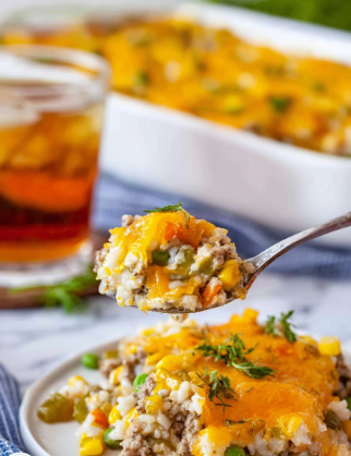 A close-up photo shows a spoon holding a scoop of a mixed dish with visible layers including yellow melted cheese, small pieces of light brown ground meat, white rice, green peas, and corn kernels, all mixed together in a creamy texture. On a white plate below the spoon, there is a serving of this dish with orange melted cheese on top and scattered green pieces, likely herbs or vegetables. In the background, there is a white baking dish filled with more of the dish topped with golden melted cheese, placed on a white marbled surface. A glass of iced tea and some green garnish are also visible behind the plate. photo taken with an iphone --ar 2:3 --v 7 - Cheesy Ground Beef and Rice Casserole, Ground Beef Rice Casserole, Easy Beef and Rice Bake, Cheesy Beef Casserole Dinner, Comfort Food Ground Beef Casserole