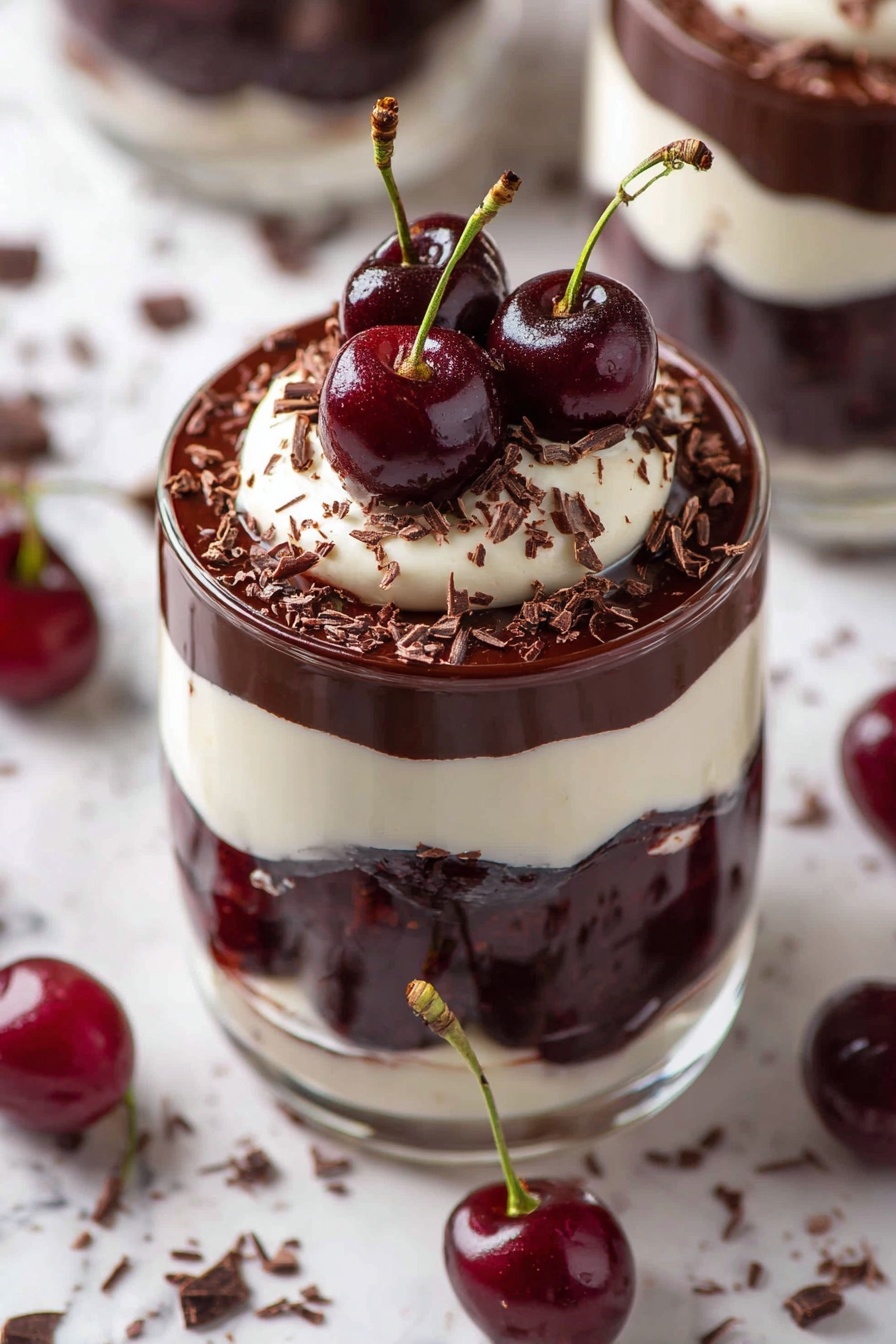 The image shows a clear glass filled with four visible layers: the bottom layer is white cream, followed by a layer of dark chocolate and whole red cherries, then another white cream layer, topped with a thick dark chocolate layer. On top, there is a dollop of white cream with three shiny dark red cherries with green stems placed on it, sprinkled with small chocolate shavings. The glass sits on a white marbled surface with some scattered chocolate shavings and cherries around photo taken with an iphone --ar 2:3 --v 7 - Vegan Black Forest Cherry Dessert Cups, vegan cherry dessert, dairy-free chocolate mousse, no-bake vegan dessert, summer fruit vegan treat