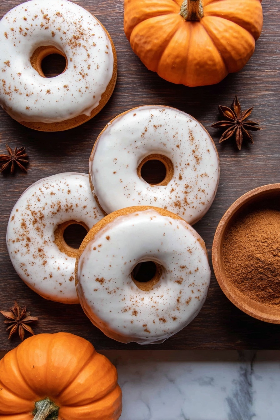 Four donuts are shown from above, each with a smooth white glaze covering the top layer, with a light dusting of brown cinnamon sprinkled unevenly on some. The donuts have a soft, golden-brown dough base visible around the hole and edges under the glaze. Three small orange pumpkins with natural ridges are placed around the donuts on a dark wooden surface. Two star anise spices sit near the top and bottom edges. On the right side, there is a small wooden bowl filled with brown powder, likely cinnamon. The background is changed to a white marbled texture. photo taken with an iphone --ar 2:3 --v 7 - Pumpkin Spice Donuts with Maple Glaze, pumpkin spice donuts, fall donut recipes, easy pumpkin donuts, homemade pumpkin donuts with glaze