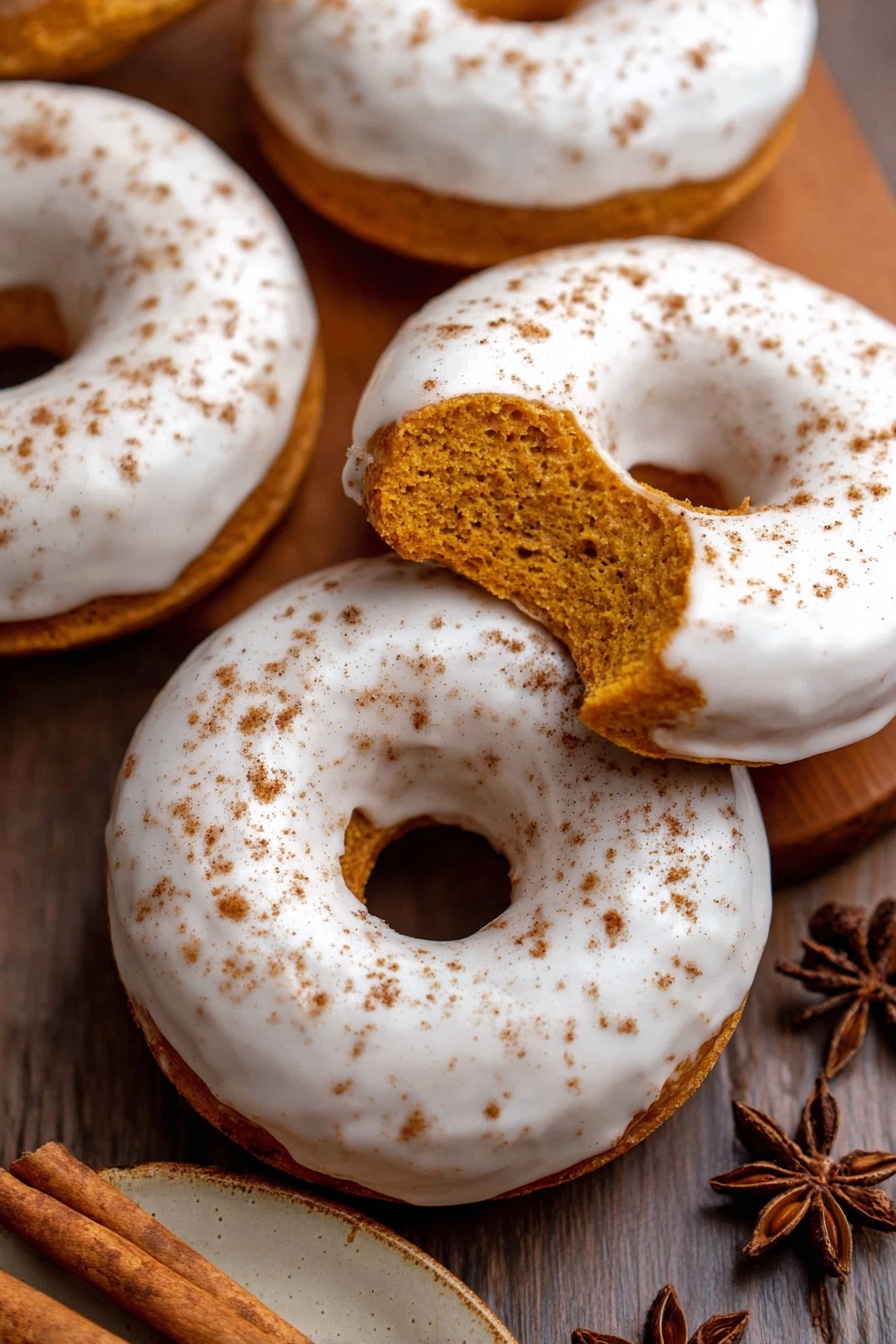 The image shows five round donuts with a smooth white icing evenly spread over the top layer, sprinkled lightly with brown cinnamon powder. One donut has a bite taken out, revealing a soft, moist orange-brown cake inside. The donuts sit on a wooden surface, and a small plate with cinnamon sticks and star anise is visible on the side. The focus is on the donuts that fill most of the frame with a cozy, warm tone. photo taken with an iphone --ar 2:3 --v 7 - Pumpkin Spice Donuts with Maple Glaze, pumpkin spice donuts, fall donut recipes, easy pumpkin donuts, homemade pumpkin donuts with glaze