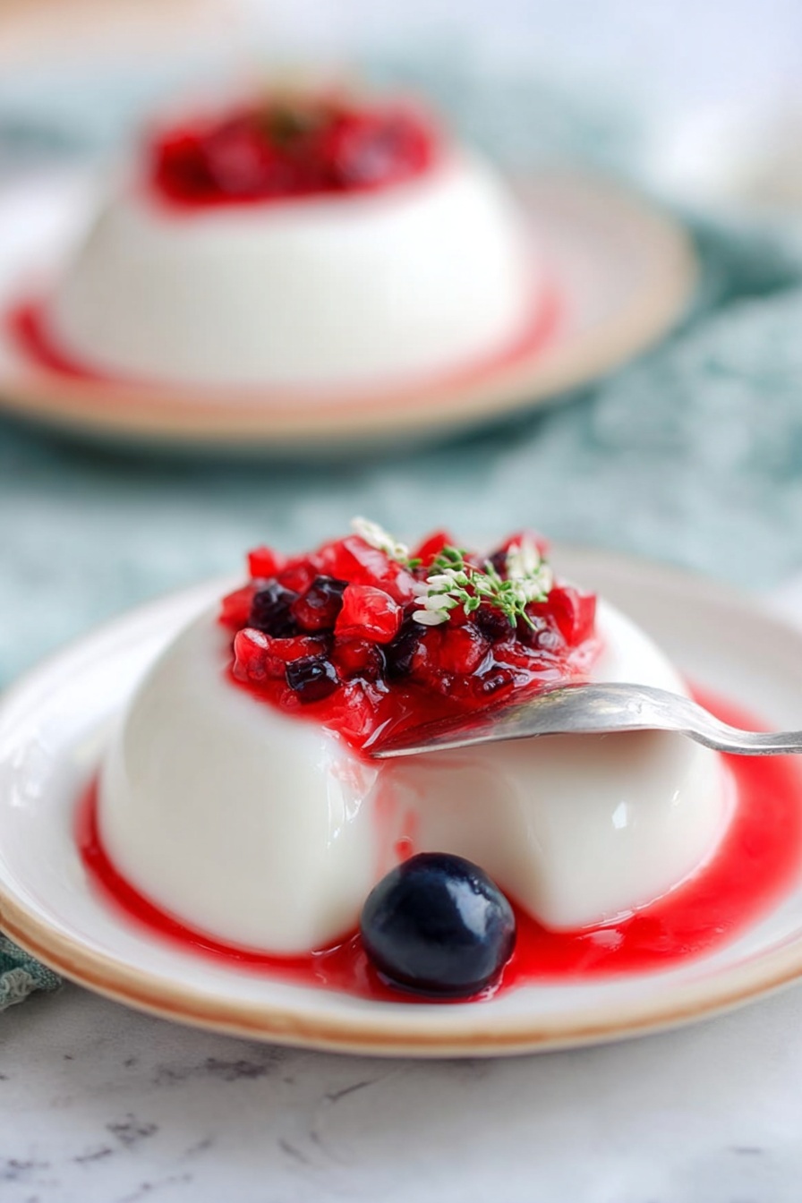 The image shows a smooth, white panna cotta dessert with one bite taken out from the front side, revealing its creamy texture. On top, there is a layer of small cubed bright red strawberries and a few whole shiny dark blue blueberries, along with a small green sprig of herb with tiny purple flowers as garnish. The panna cotta sits on a white plate with a green center and a thin golden rim. The plate is on a white marbled surface with soft, blurred pastel colors in the background. Photo taken with an iphone --ar 2:3 --v 7 - Greek Yogurt Panna Cotta with Thyme Berries, Greek yogurt dessert, easy panna cotta recipes, berry and thyme dessert, light summer desserts