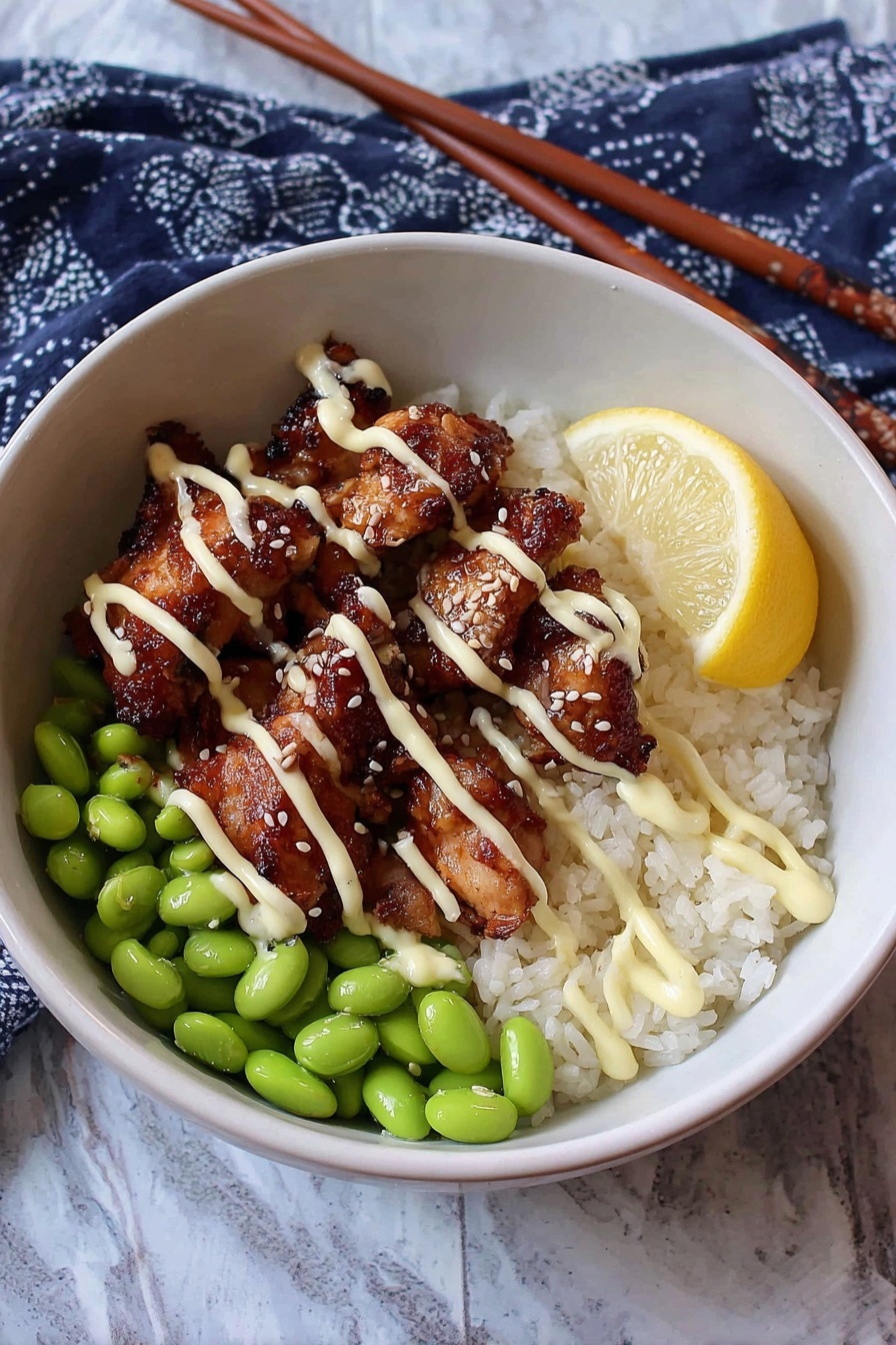 A white bowl holds a meal with three main layers: the bottom layer is white rice with a slightly sticky texture, the middle layer has bright green edamame beans arranged neatly on one side, and the top layer consists of several pieces of dark brown cooked chicken with a shiny glaze. Light cream-colored sauce is drizzled over the chicken in thin lines, and small white sesame seeds are sprinkled on top. A yellow lemon wedge rests at the edge of the bowl. The bowl sits on a white marbled surface with a dark blue cloth with white patterns nearby, and there are brown chopsticks to the side. Photo taken with an iphone --ar 2:3 --v 7 - Air Fryer Karaage Chicken, Japanese fried chicken, crispy chicken recipe, healthy fried chicken, easy air fryer chicken