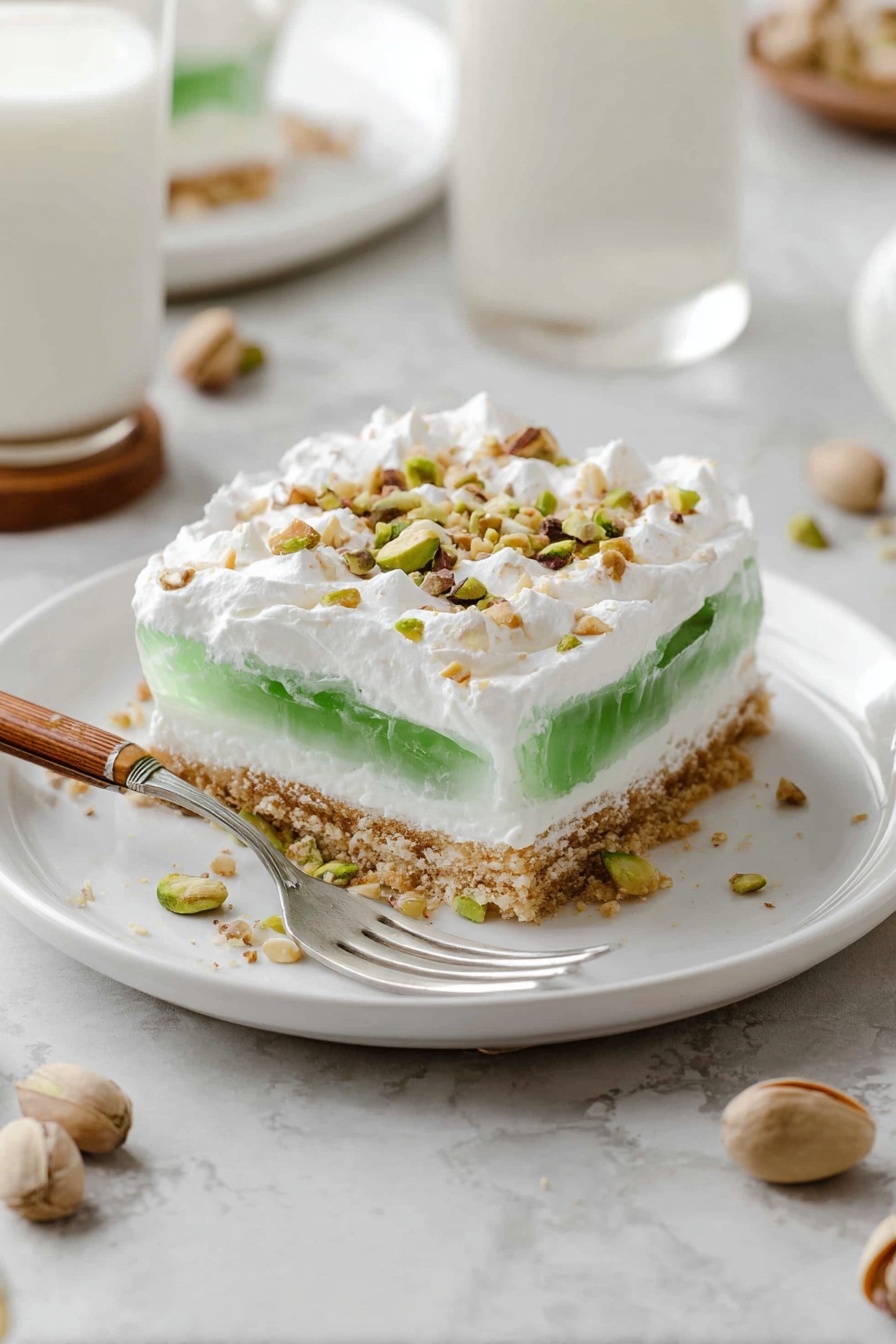 A white plate holds a square piece of layered dessert with four layers: a light brown crumbly base, a thick white creamy layer above it, a green jelly-like layer in the middle, and a thick white whipped topping on top sprinkled with small pieces of nuts. A fork with a wooden handle is placed on the plate, slightly under the dessert with some whipped topping on the fork. The plate rests on a white marbled surface, with scattered pistachio shells nearby. In the background, there is a blurred glass of milk on a coaster and a white plate with the remaining dessert. photo taken with an iphone --ar 2:3 --v 7 - Layered Pistachio Dessert, pistachio dessert, no-bake pistachio treat, festive pistachio layered dessert, easy pistachio layered dessert