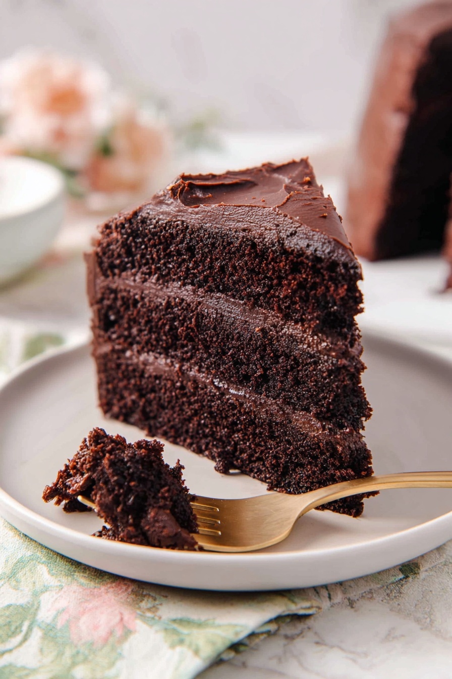 A moist chocolate cake slice with three thick layers, each dark brown and rich in texture, separated by smooth and creamy chocolate frosting layers. The outside is covered with the same dark chocolate frosting, slightly uneven but thickly spread. A small piece of the cake is held on a gold fork in the front, showing the crumbly, soft texture inside. The cake sits on a white plate, placed on a white marbled surface with a soft floral cloth nearby. photo taken with an iphone --ar 2:3 --v 7 - Ultimate Chocolate Cake, moist chocolate cake, rich chocolate dessert, homemade chocolate cake, easy chocolate cake recipe