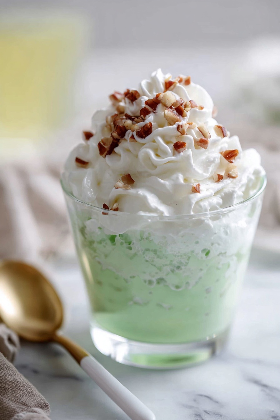 The image shows a clear glass filled with two layers: the bottom layer is bright light green with a creamy texture and small bits mixed inside, while the top layer is a thick swirl of white whipped cream, sprinkled with small pieces of chopped brown nuts. The glass is placed on a white marbled surface, and in the background, there is an out-of-focus light yellow drink. A spoon with a white handle and gold tip lies next to the glass. photo taken with an iphone --ar 2:3 --v 7 - Easy Pistachio Watergate Salad, Pistachio Watergate Salad, Watergate Salad with Pistachios, No-Bake Watergate Salad, Retro Pistachio Fruit Salad